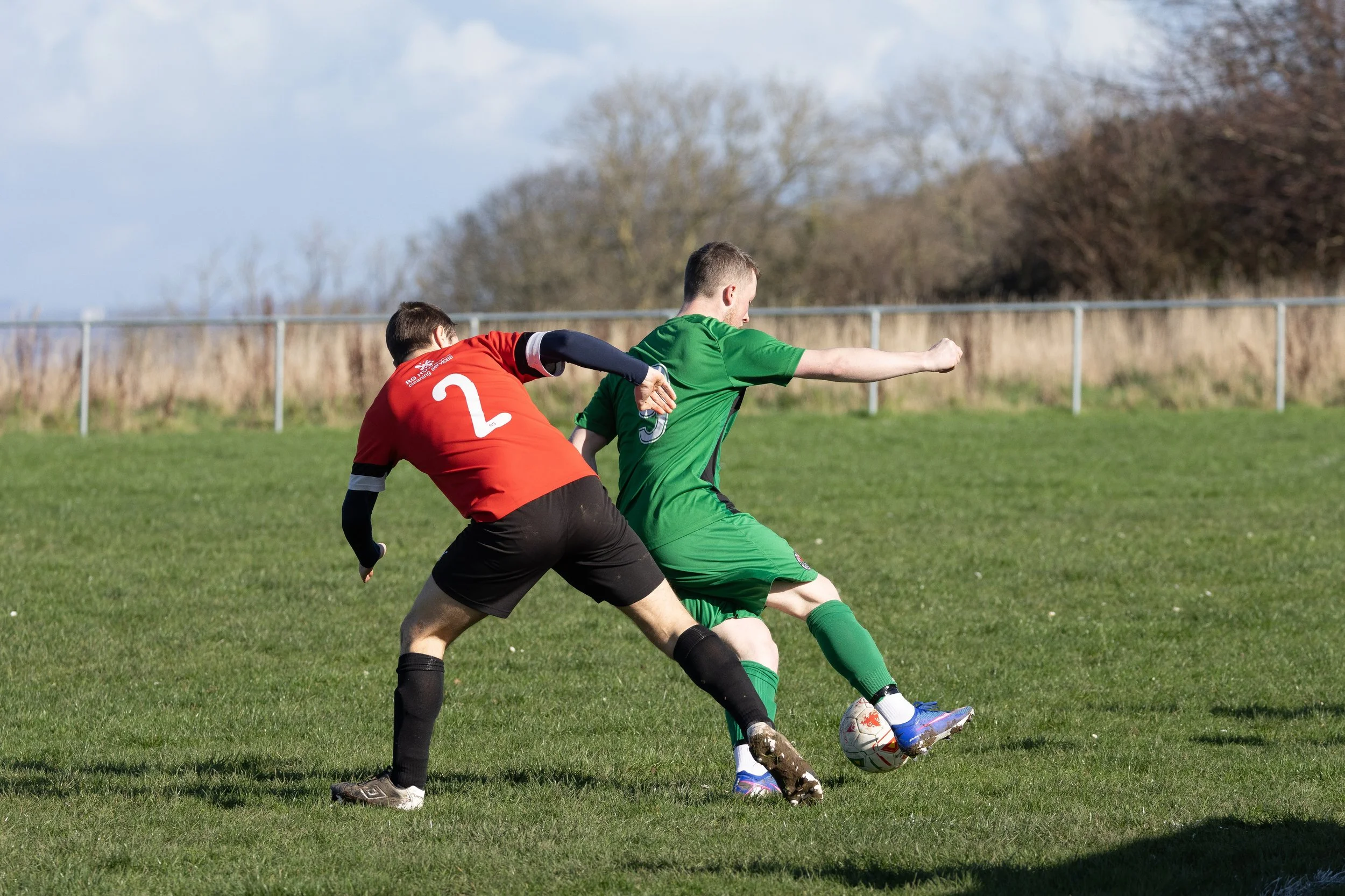 Two male soccer players competing for the ball on a grassy field, one in a red jersey and the other in a green jersey, with trees and a fence in the background.