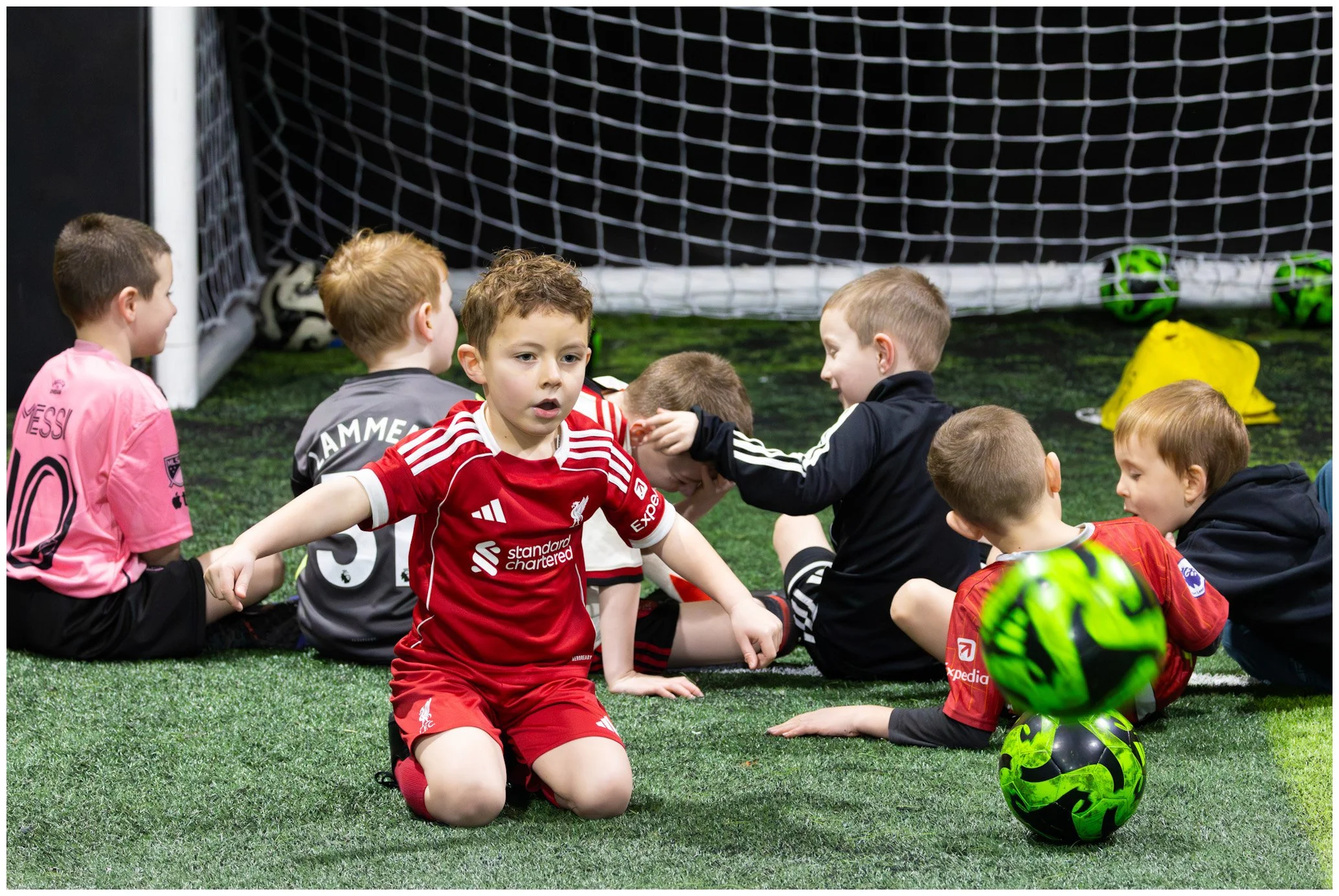 Young boys sitting on artificial turf inside an indoor soccer facility, some wearing soccer jerseys, and a goal with soccer balls in the background.