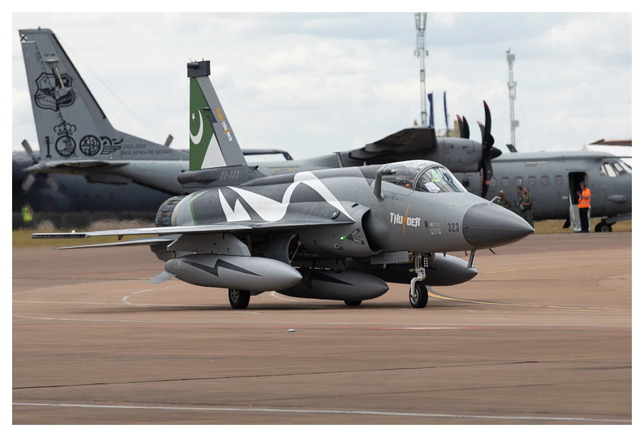 A fighter jet on an airport runway with two larger aircraft and a group of people in the background.