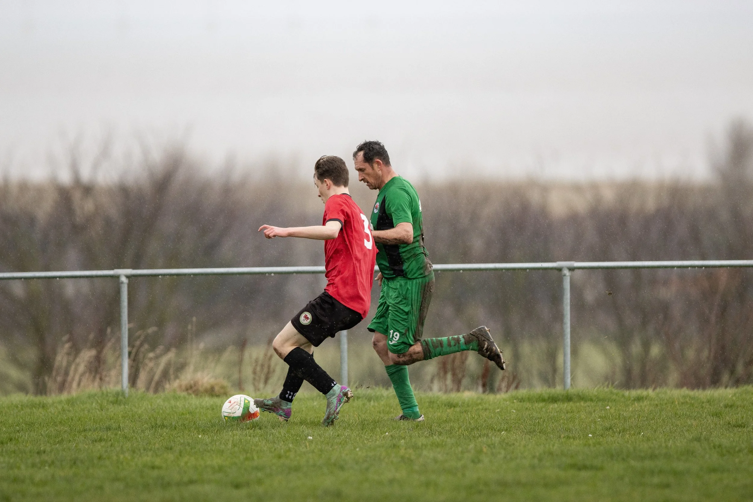 Two soccer players, one in red and the other in green, competing for the ball on a grassy field under rainy weather.
