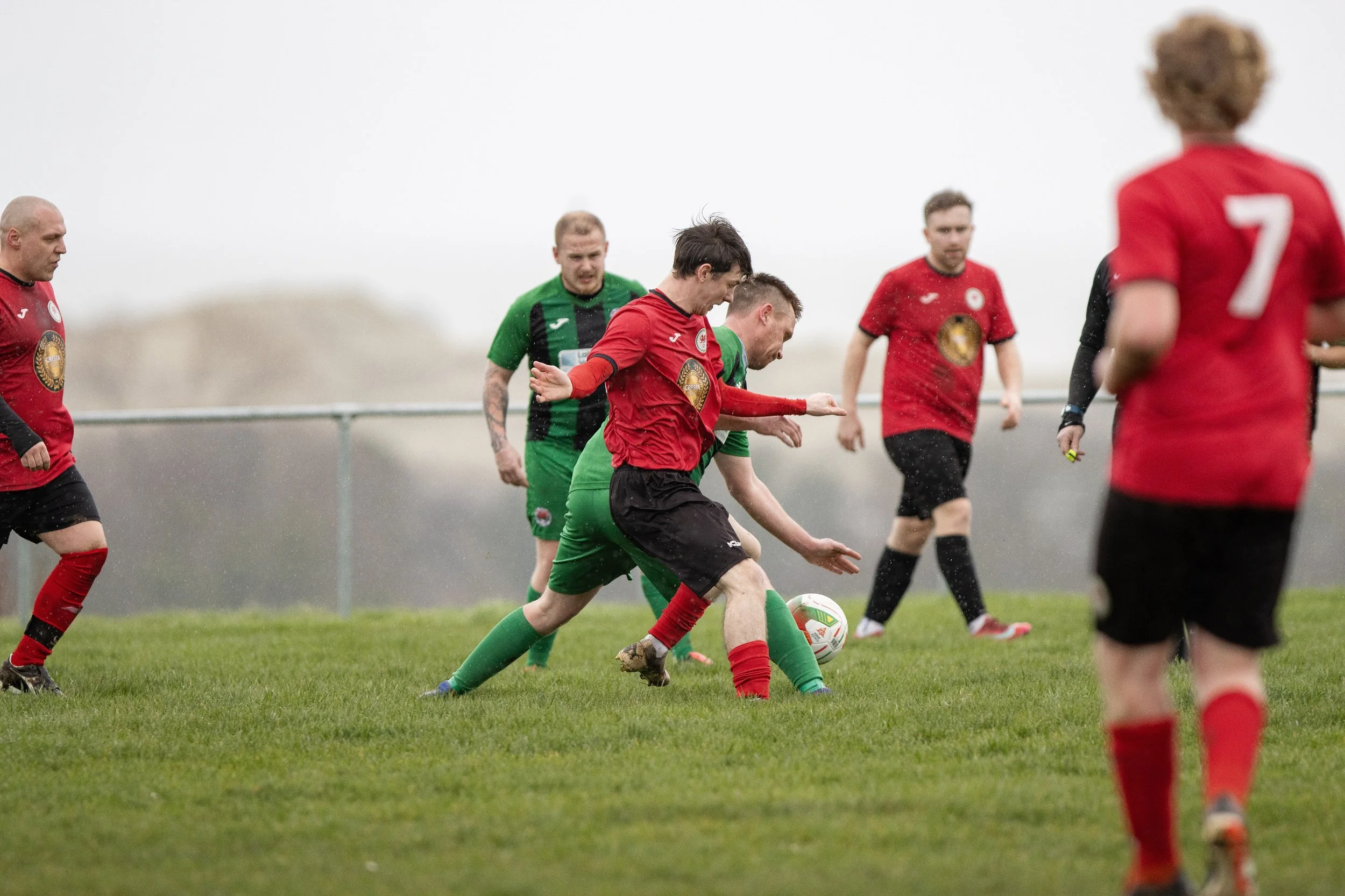 Soccer match on a rainy day with players in red and green jerseys competing for the ball on a grassy field.