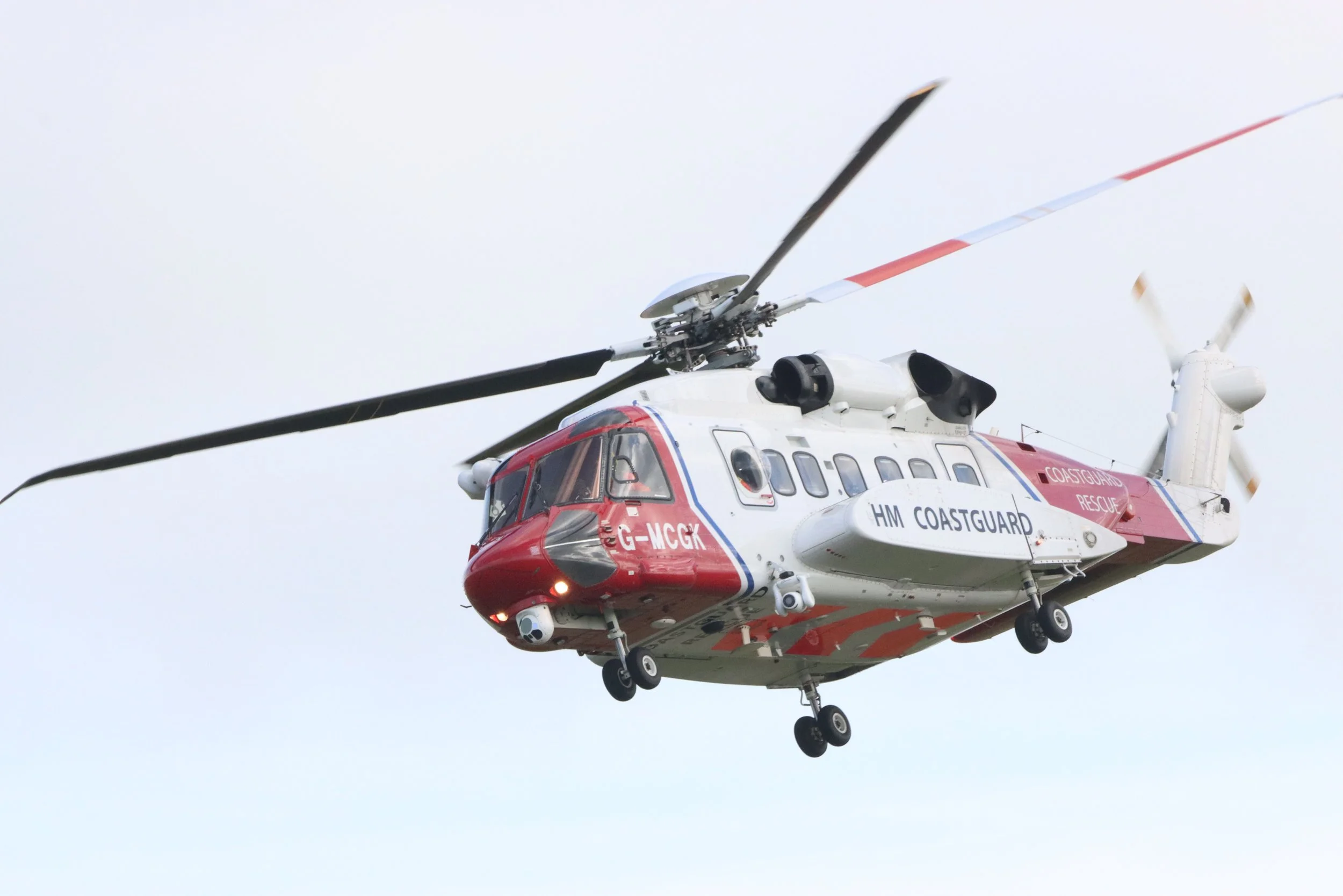 A helicopter with red and white markings labeled HM Coastguard, flying against a pale sky.