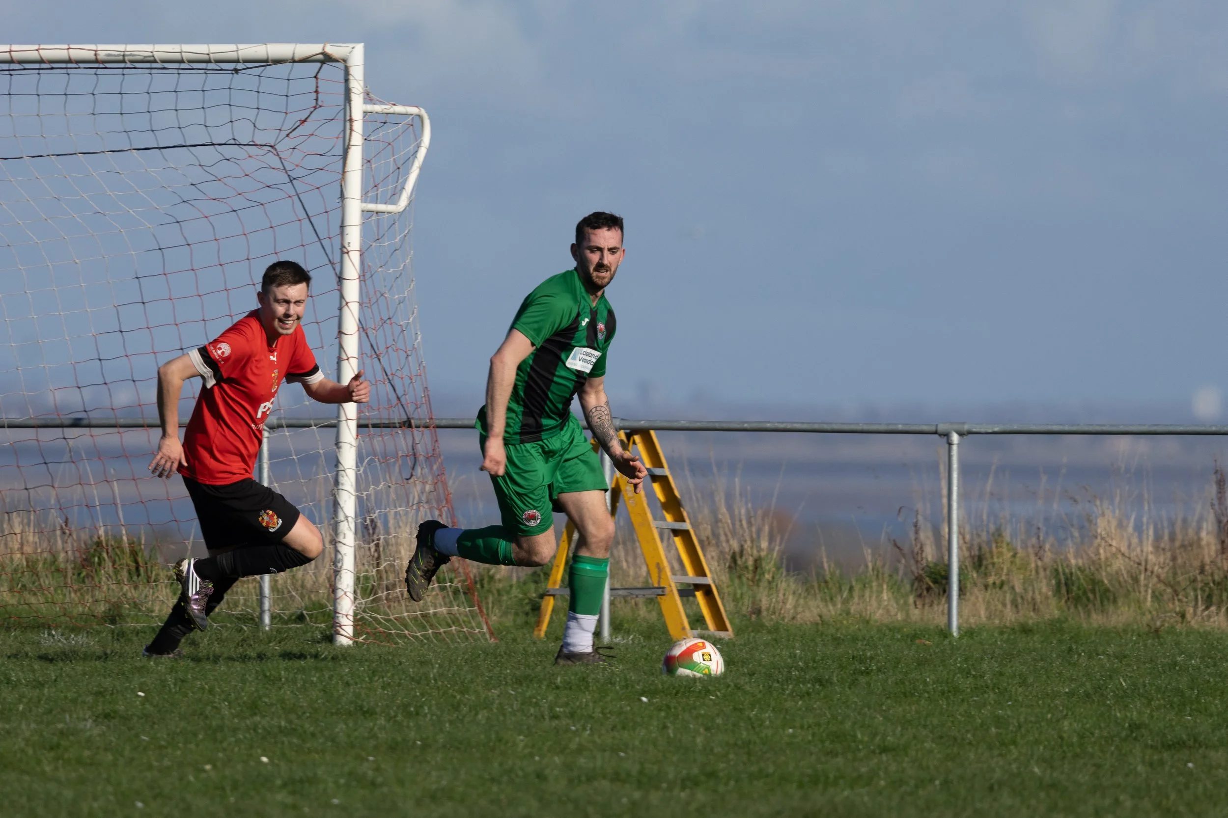 Two soccer players, one in a red jersey and the other in a green jersey, near the goalpost during a soccer match on a grassy field.