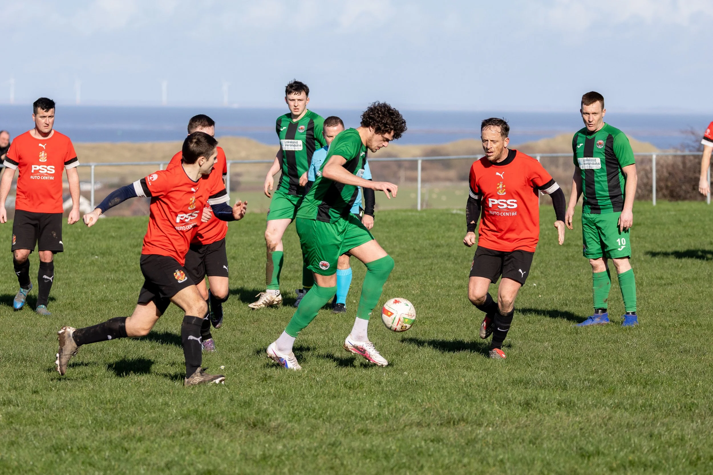 Soccer match with players in red and green jerseys on a grass field near the coast during daytime.