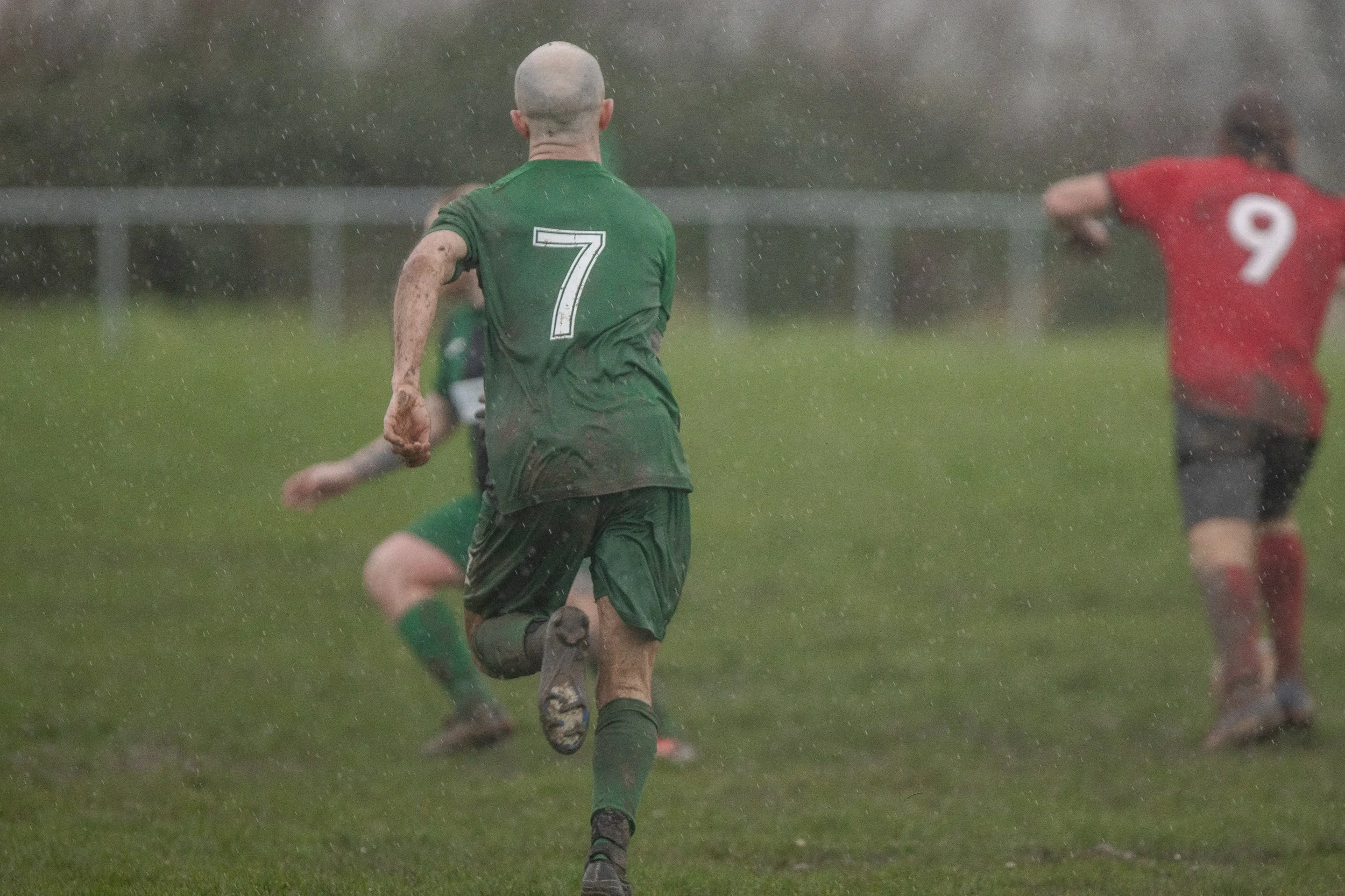 Soccer players running on a wet field during a game, with one player in a green jersey with the number 7 visible from the back, and other players in red jerseys in the background.