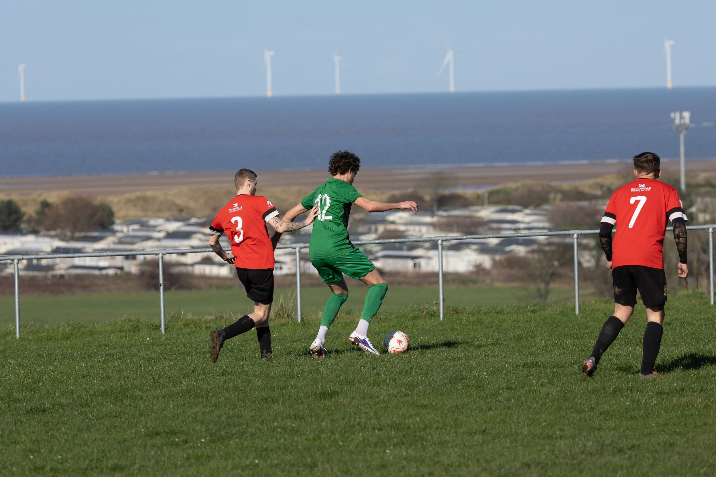 Three soccer players on a grassy field, two in red jerseys and one in green, with a scenic backdrop of water, wind turbines, and distant buildings.