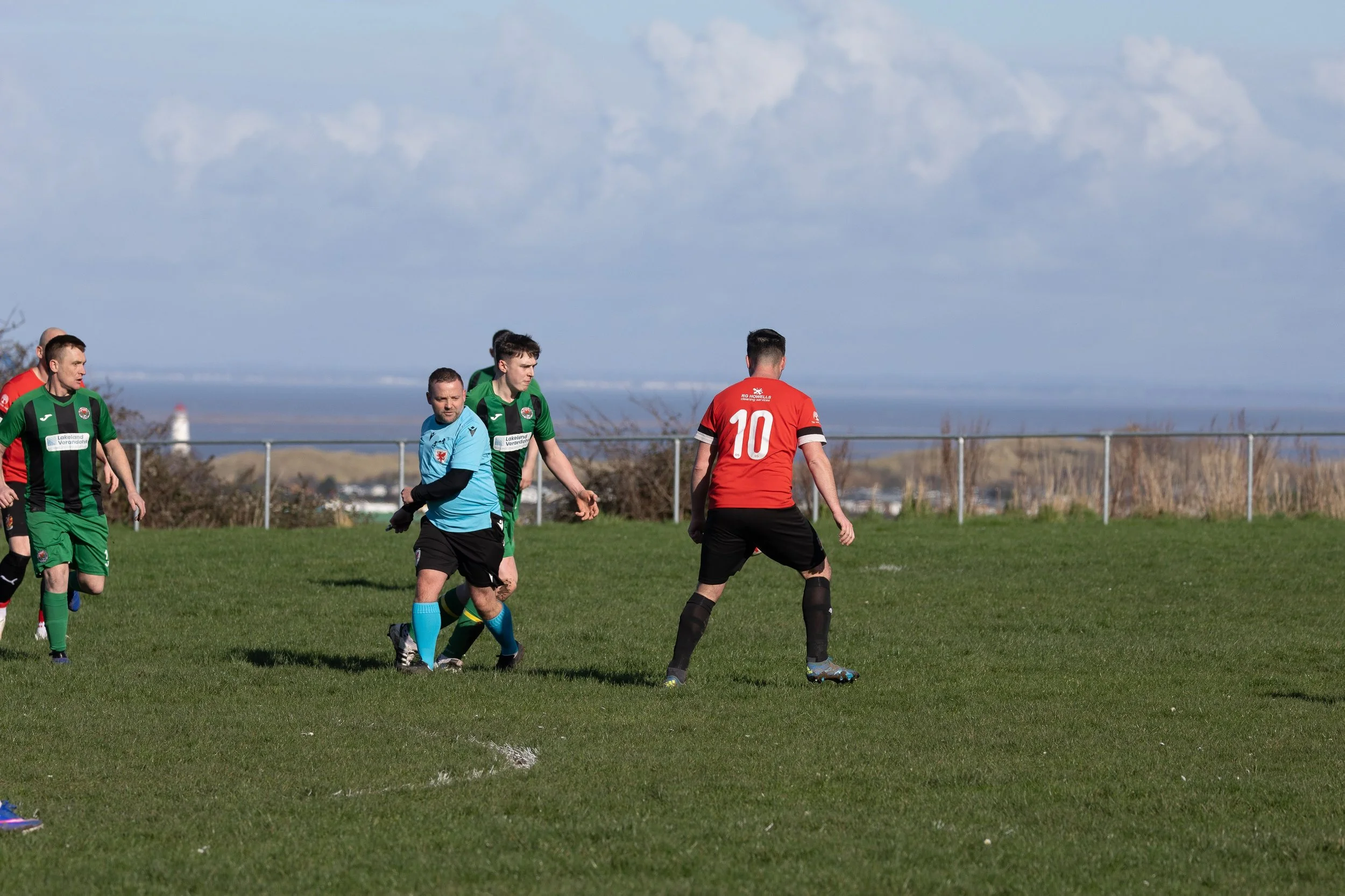 Soccer players on a field, one in a red jersey with the number 10, others in green and black uniforms, with a scenic background of hills, a lighthouse, and cloudy sky.