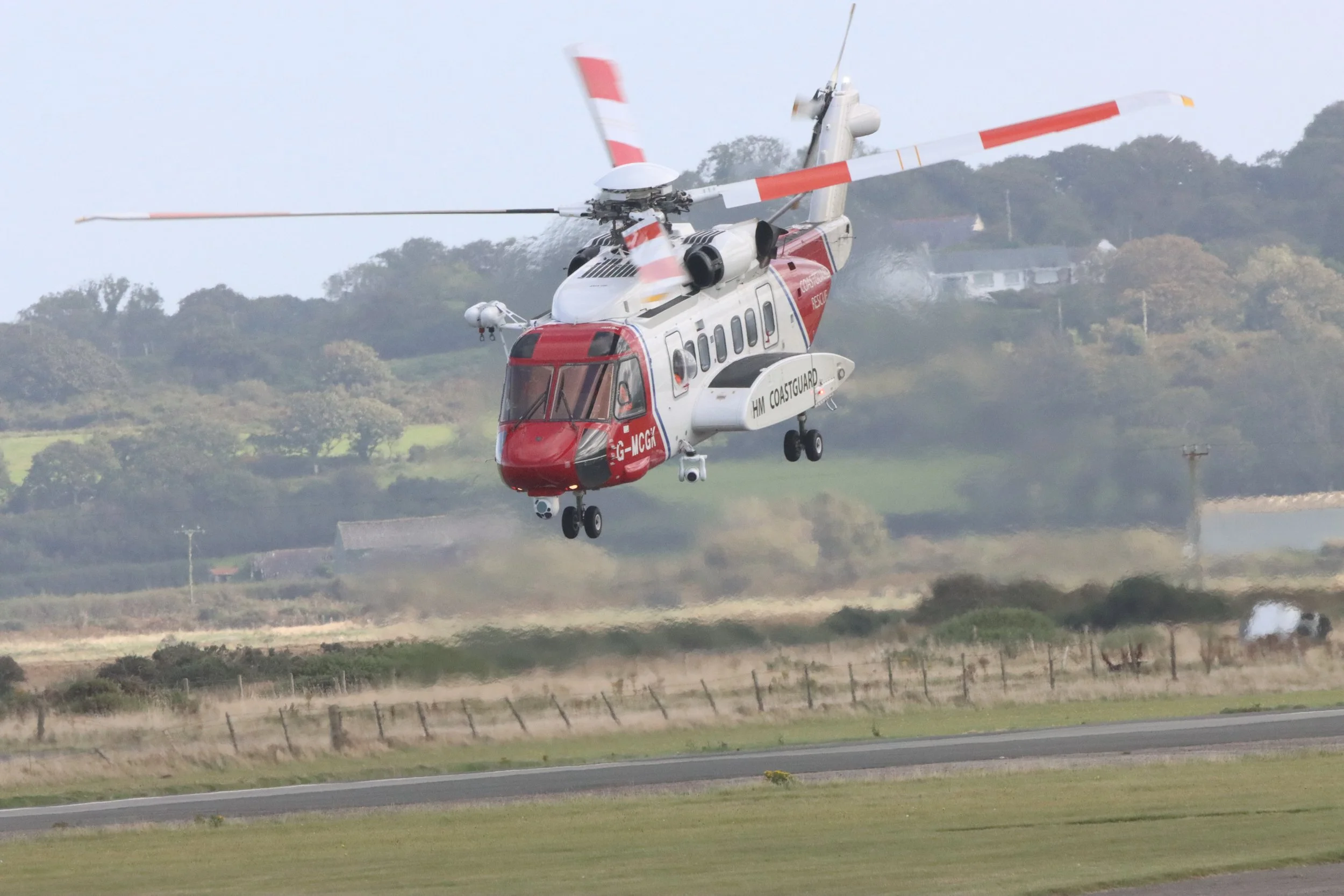 A red and white helicopter in mid-flight over a grassy field, with trees and hills in the background.