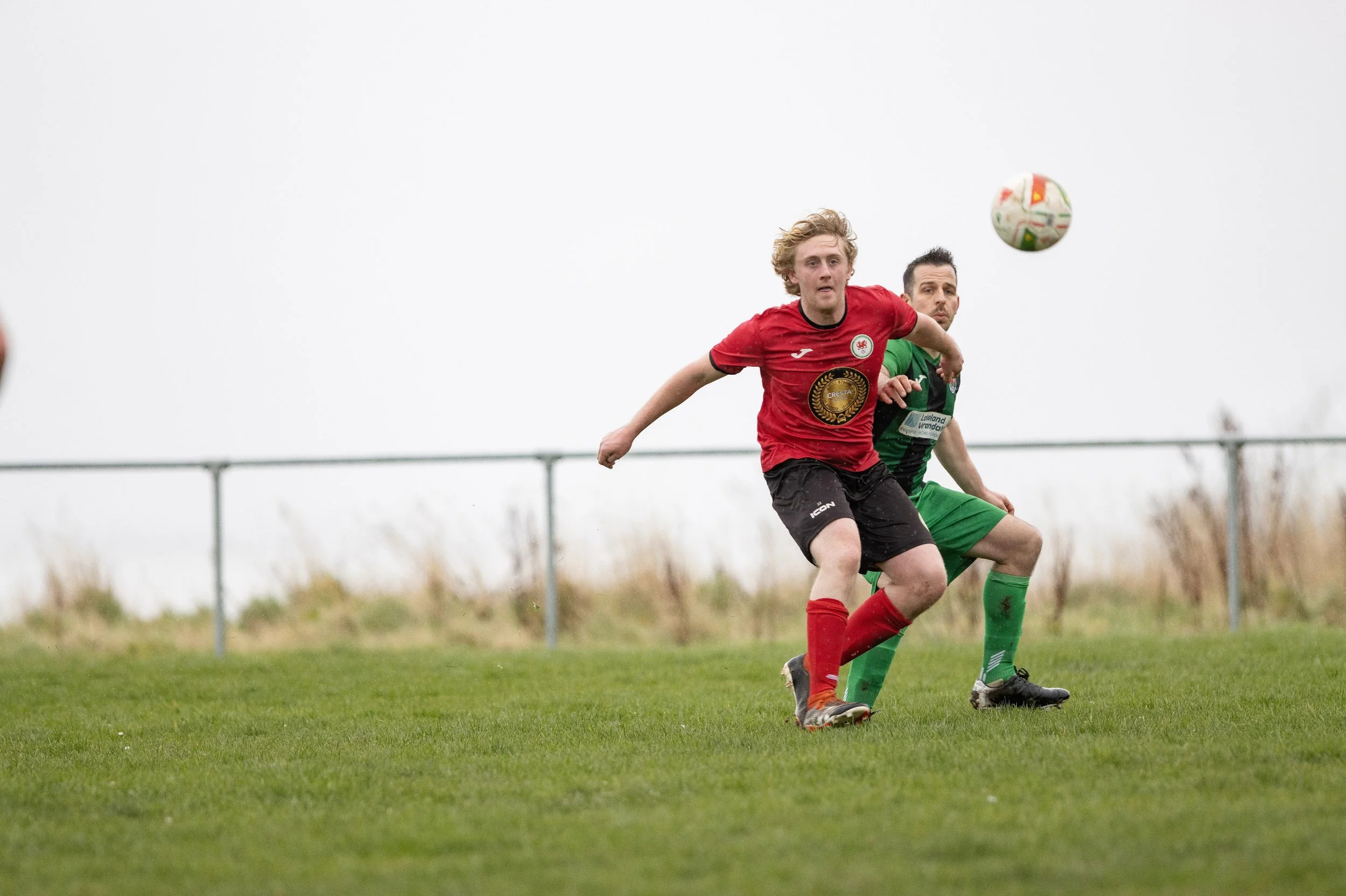 Two soccer players running after a ball on a grassy field, one in a red jersey and black shorts, the other in a green jersey and shorts, with overcast sky in background.