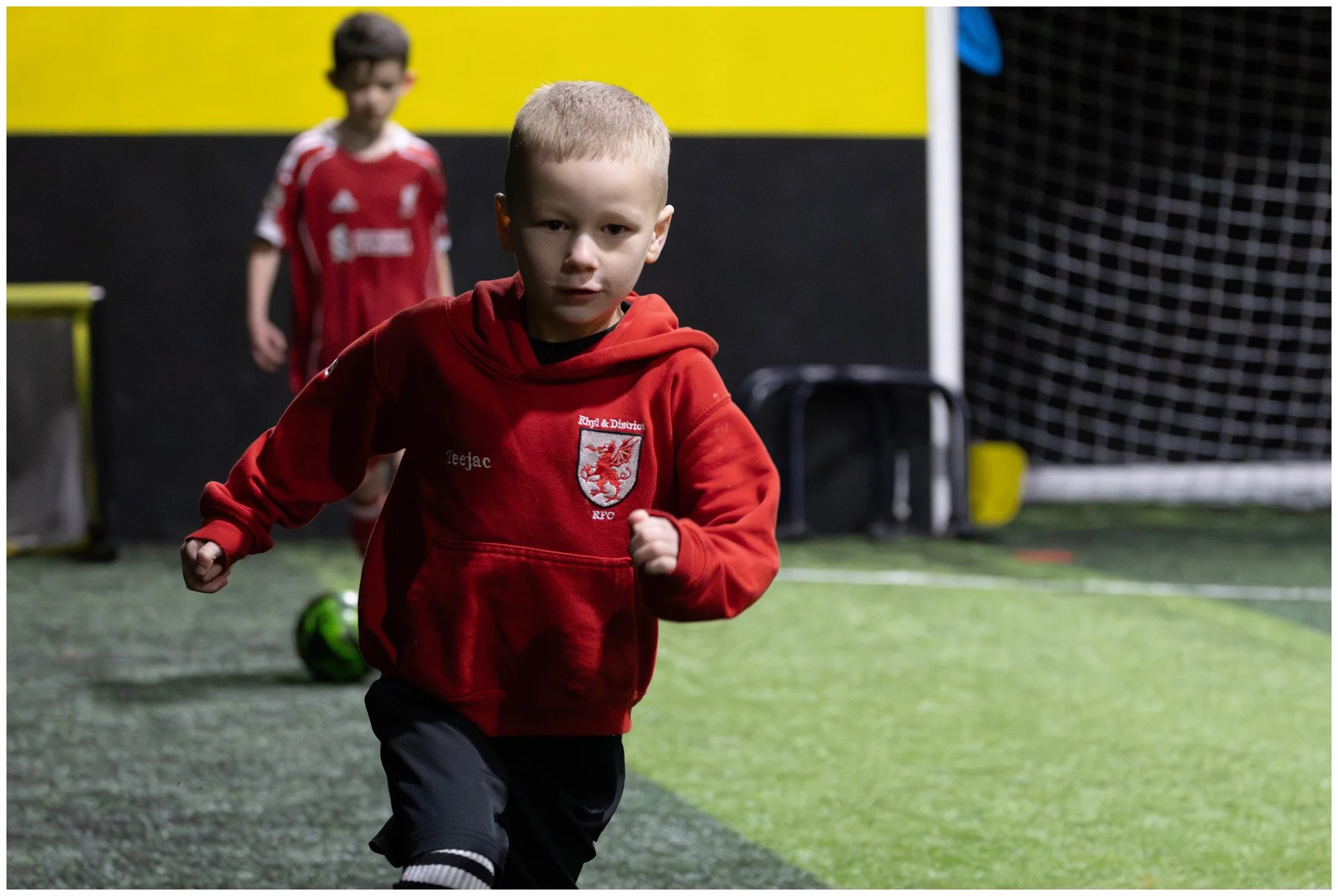 Young boy in a red hooded sweatshirt running on indoor artificial turf with another child in red jersey in the background.
