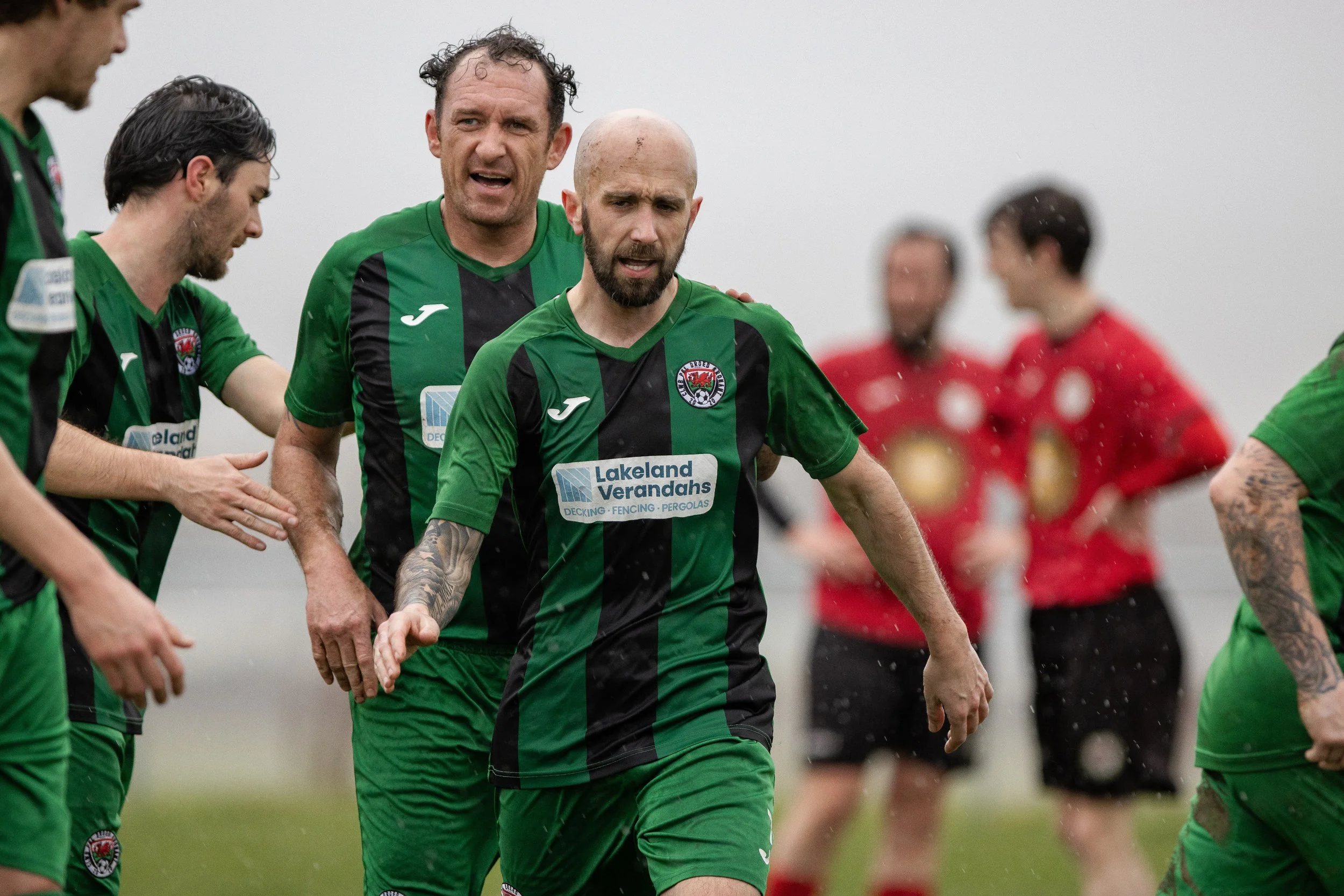 Soccer players on the field, wearing green and black jerseys, with one player in the foreground walking, and others in the background, amid rain, with a couple of players in red jerseys also visible.