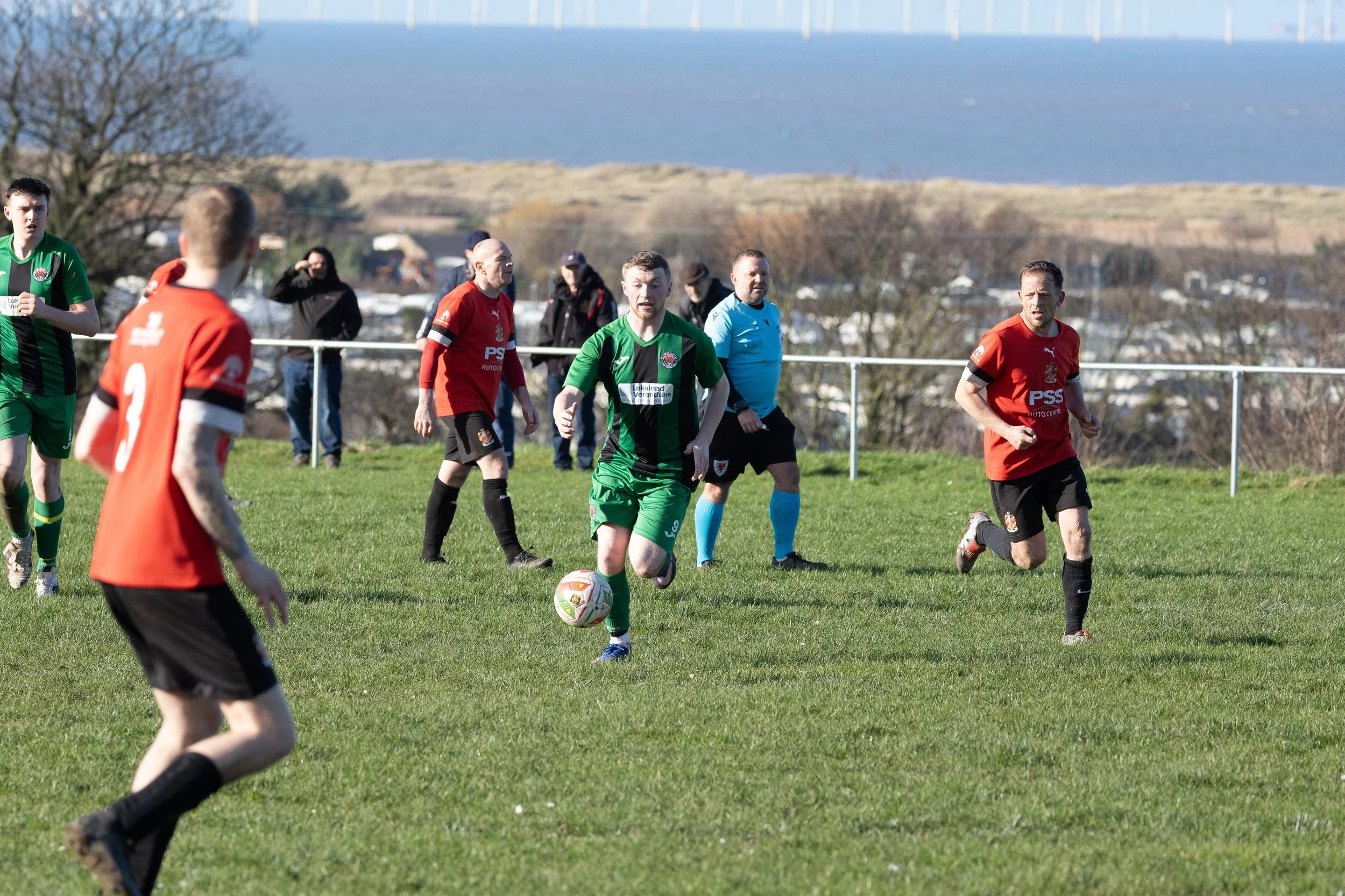 Soccer match on a grassy field with players wearing red and green jerseys. One player in a green jersey is kicking the ball, while others watch or run. A referee in a blue shirt and black shorts stands in the background. Some spectators are seen behi