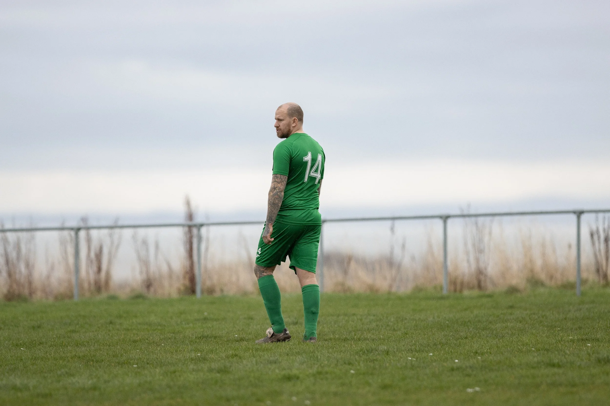 A soccer player in a green uniform with the number 14 on the back, standing alone on a grassy field, looking to his right, with a cloudy sky overhead.
