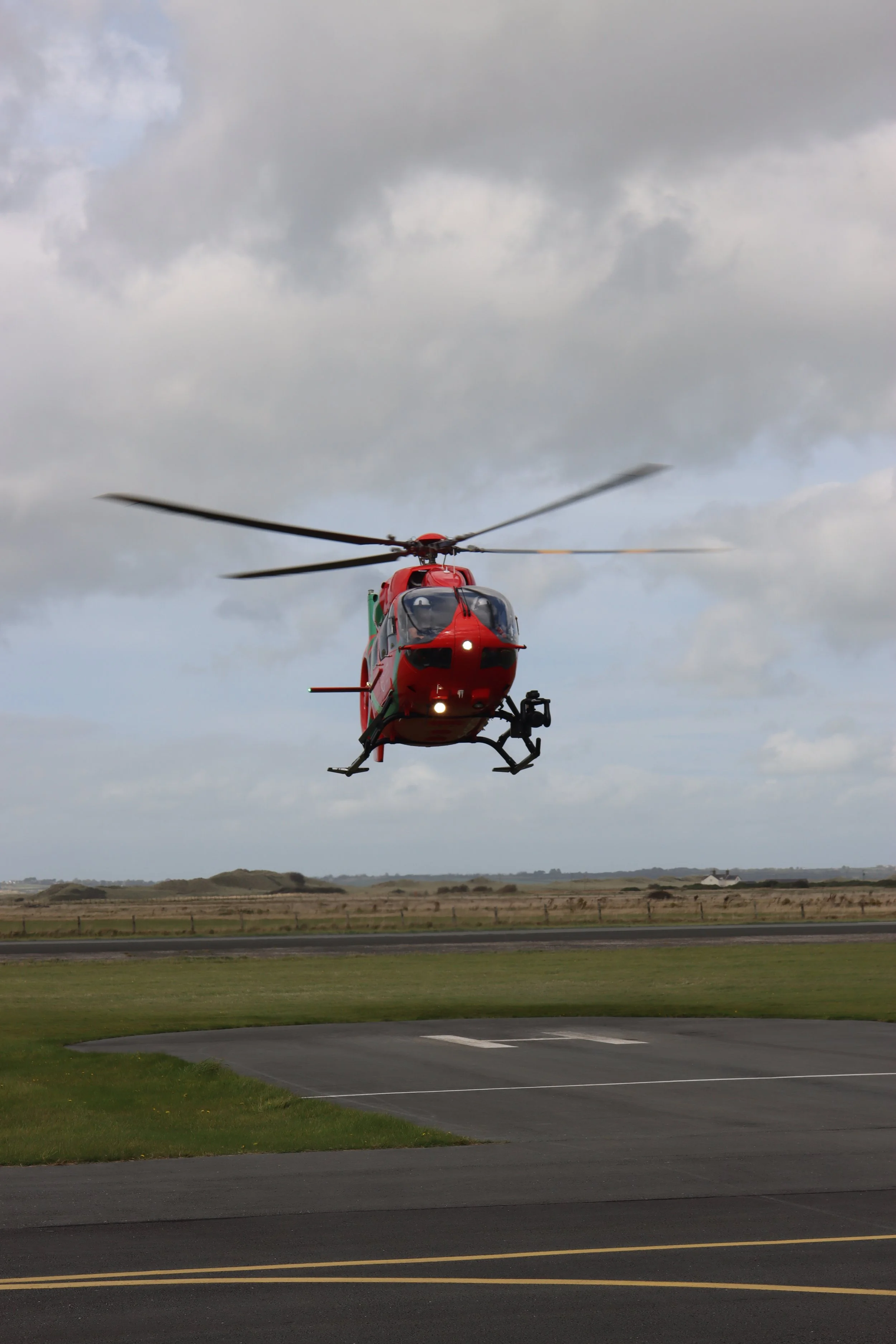 A red helicopter flying low above a paved airport tarmac with grassy fields and cloudy sky in the background.