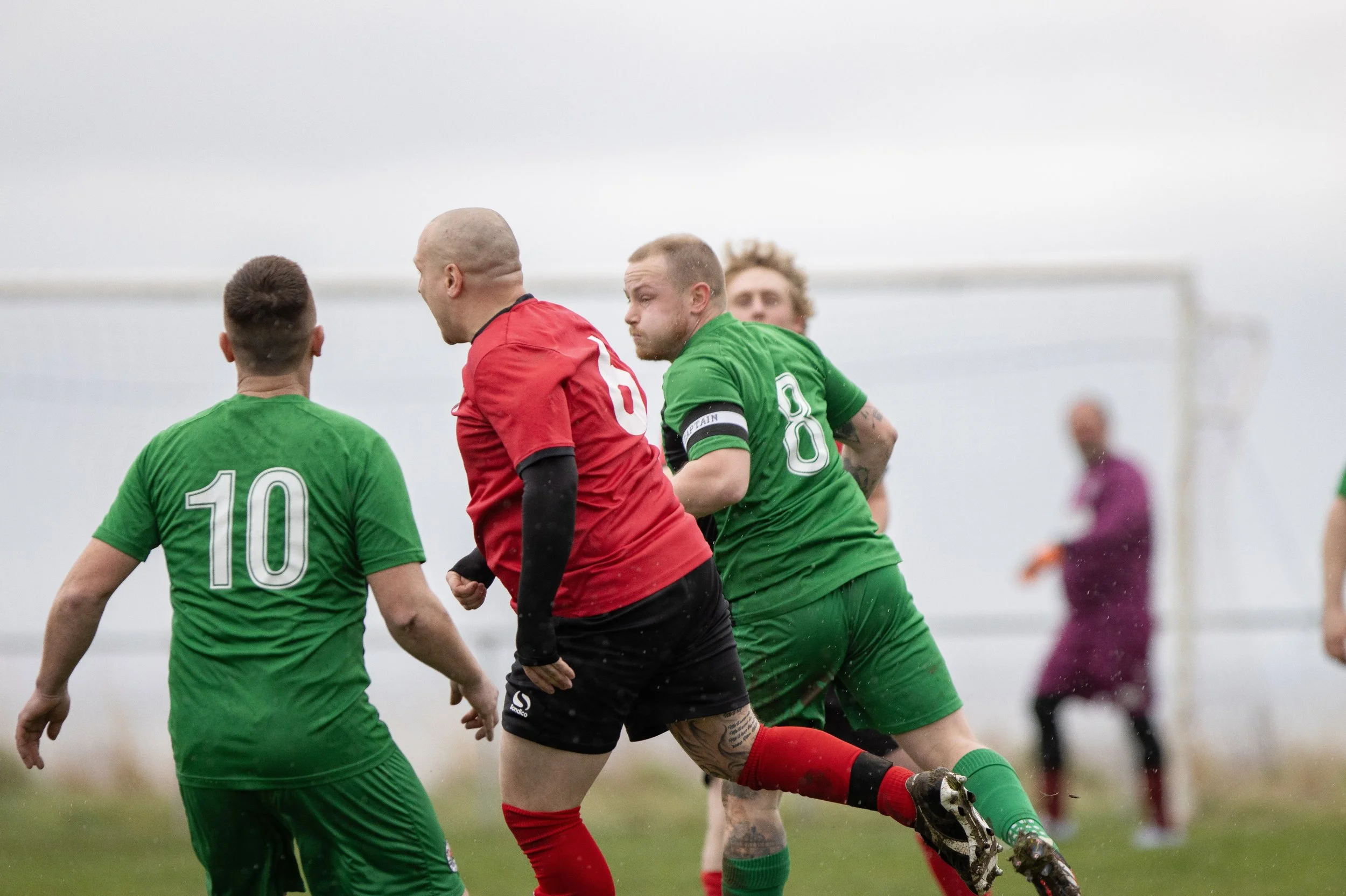 Soccer players in green and red uniforms competing for the ball on the field, with a goalkeeper in a purple kit in the background.