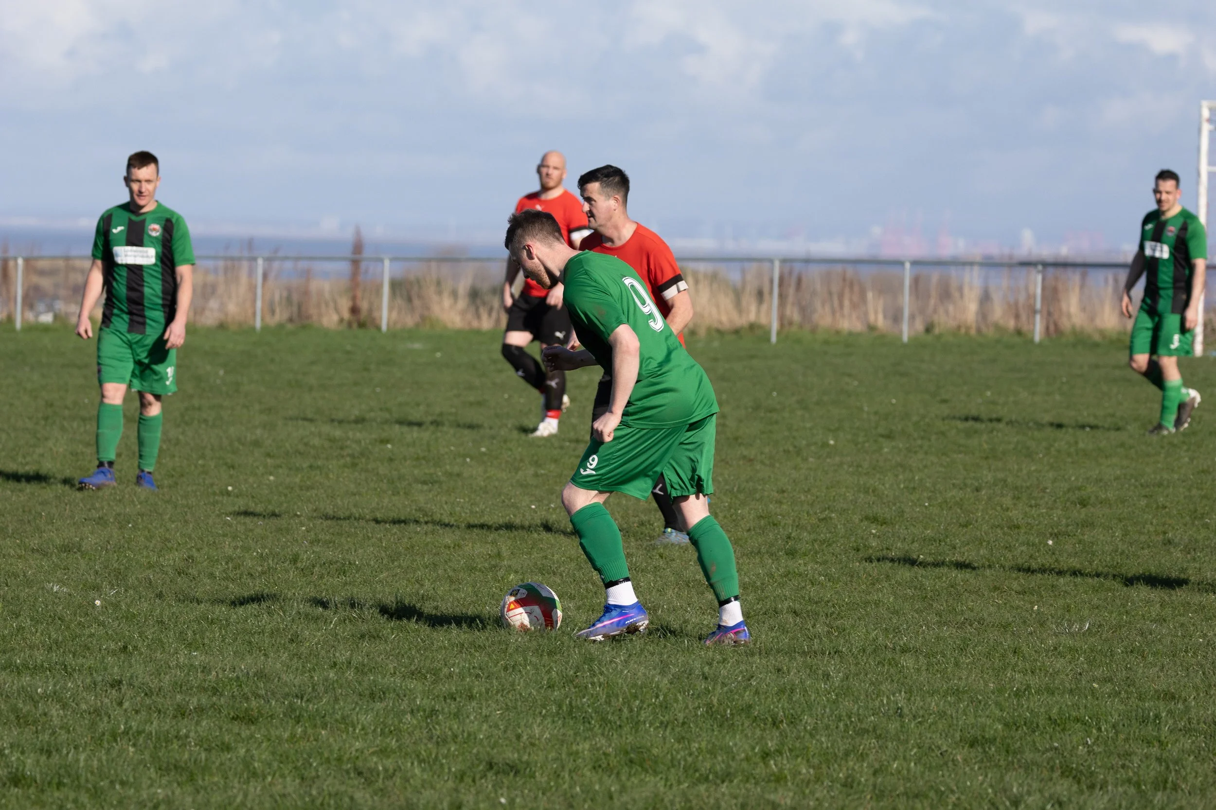 Soccer players on the field, one in green with the number 9, preparing to kick the ball, with other players in the background during a match.
