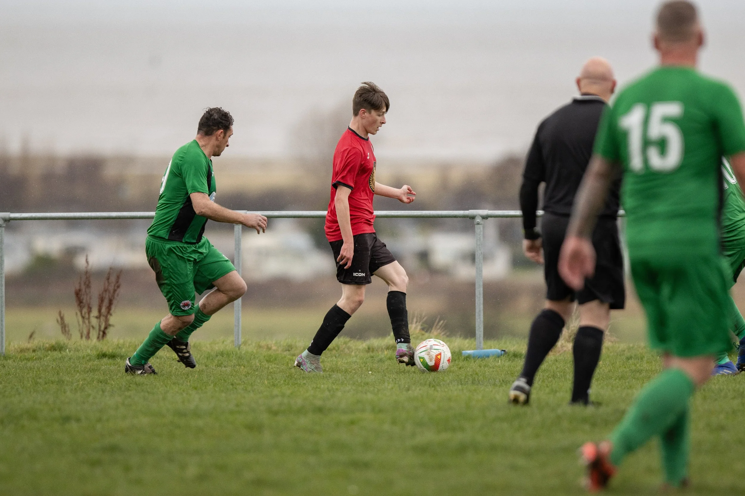 Soccer players in green and red jerseys playing on a grassy field with a player in a red jersey preparing to kick the ball, on a cloudy day.