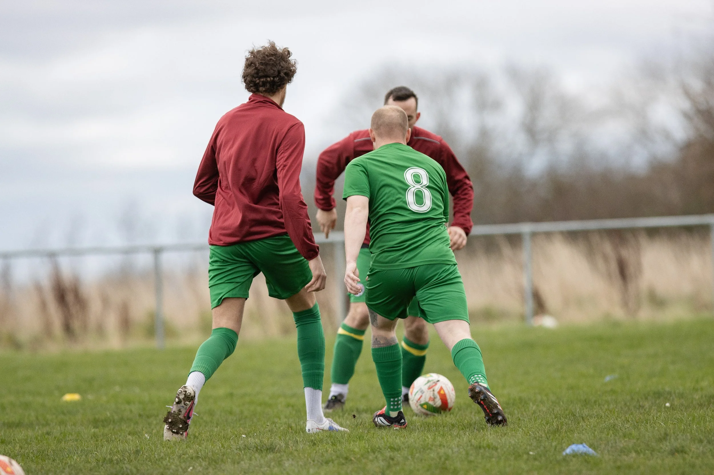 Three soccer players on a field, two in red jackets and green shorts, one in a green jersey with the number 8, competing for the ball.
