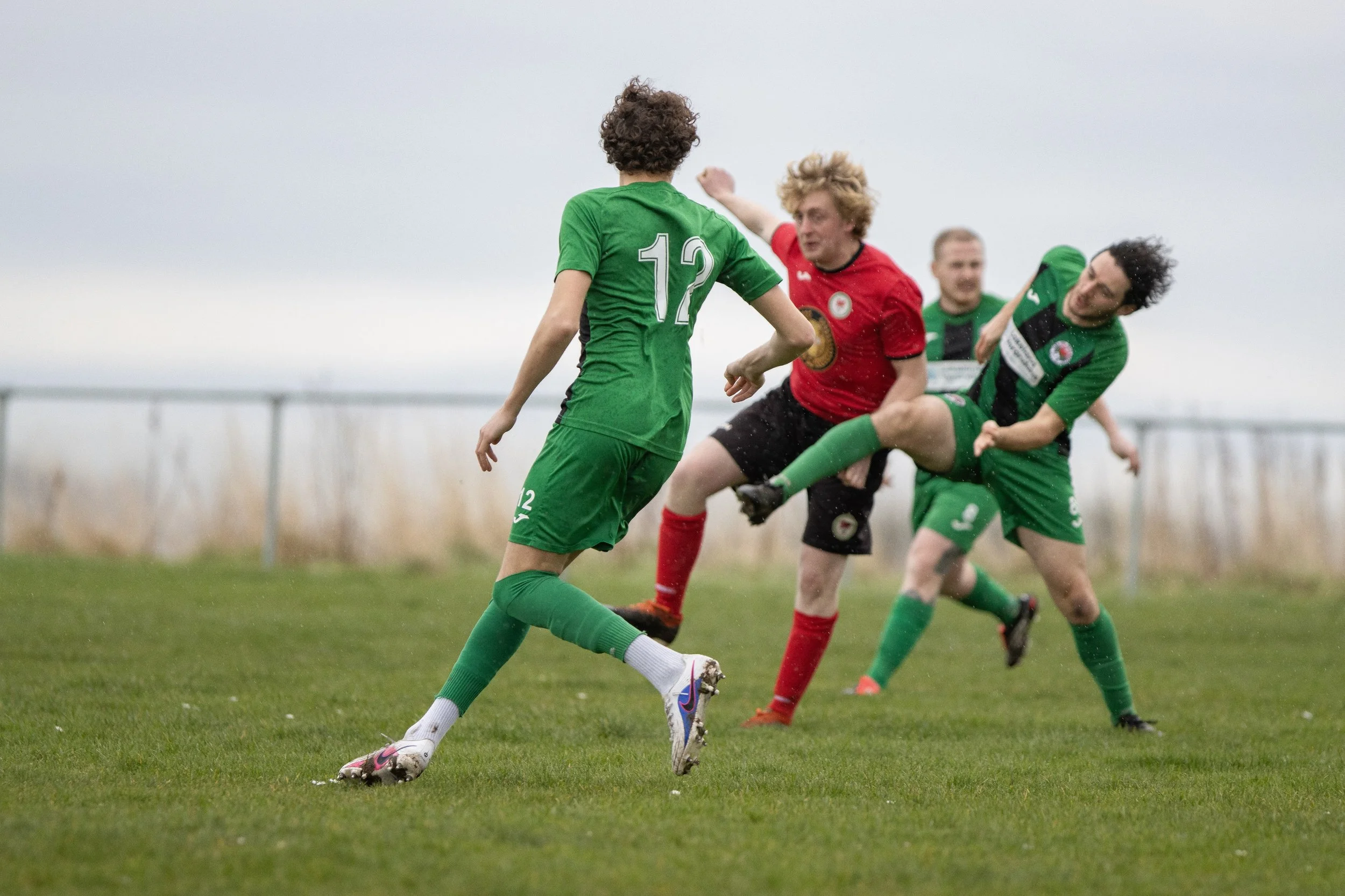 Soccer players competing for ball on grassy field, with two players in green jerseys and one player in a red jersey, during daytime.