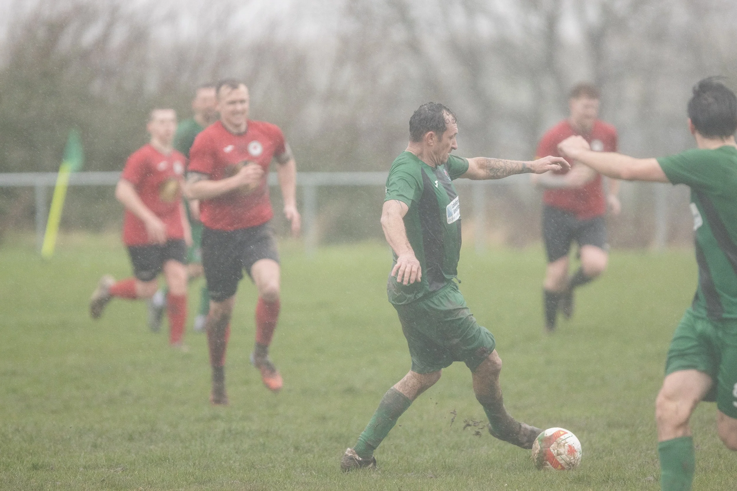 Soccer players playing in the rain on a grassy field, with one player in green controlling the ball and others in red running in the background.