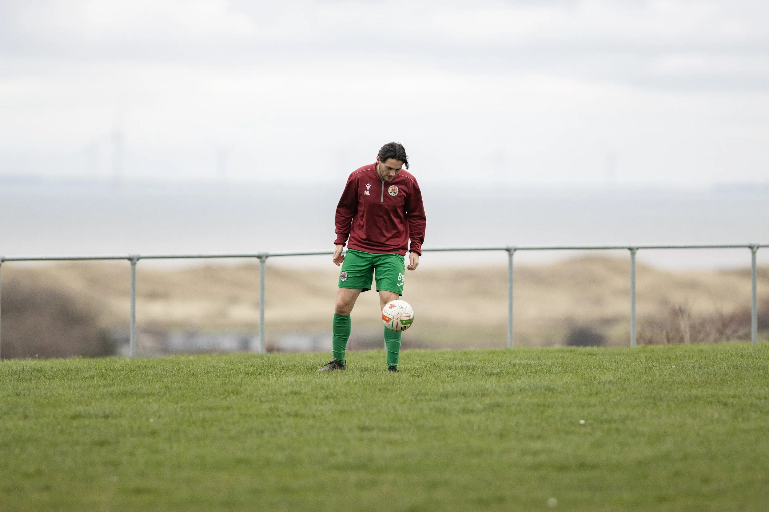 A soccer player in a maroon jacket and green shorts balancing a soccer ball on his right foot on a grassy field.
