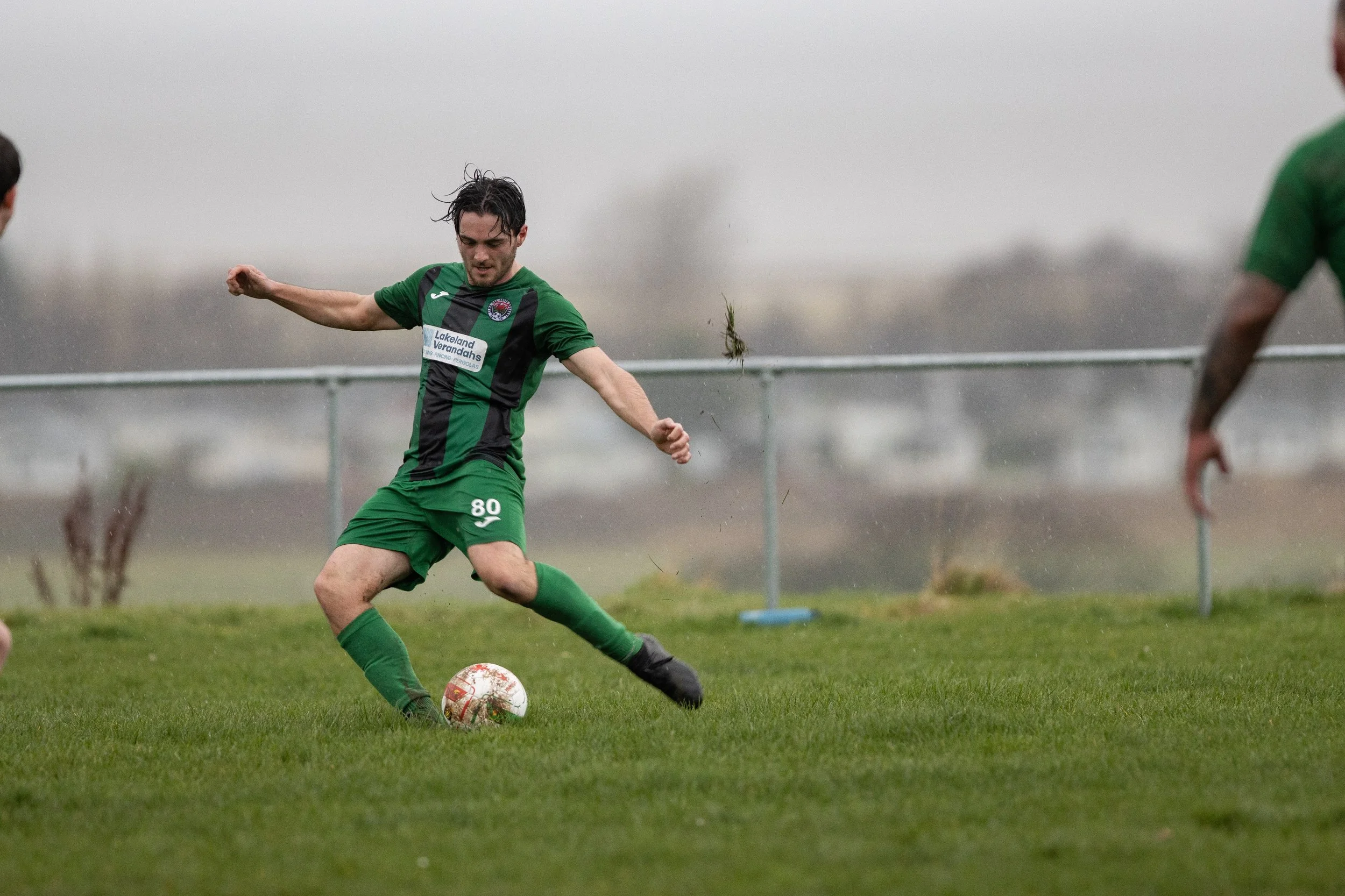 A soccer player in a green and black uniform kicking a ball on a grassy field in rainy weather.