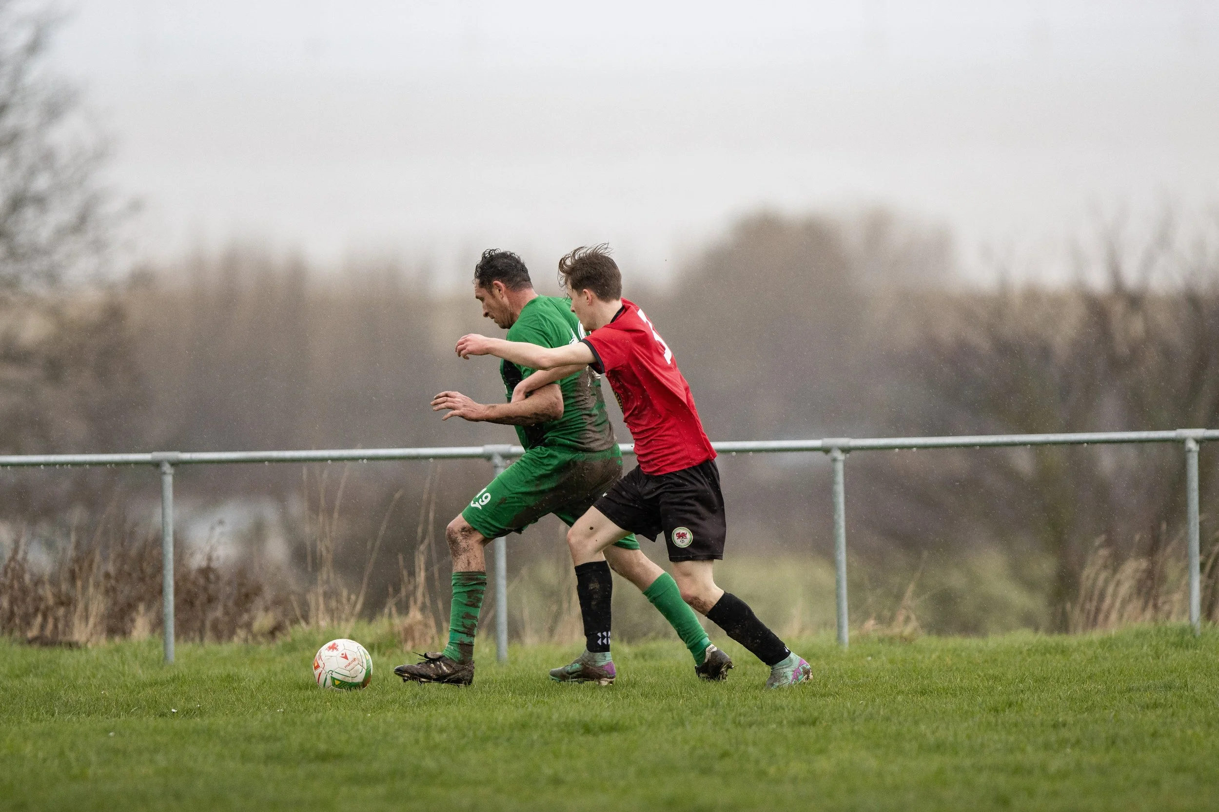 Two soccer players, one in green and one in red, competing for the ball on a grassy field with a blurry background of trees and a gray sky.