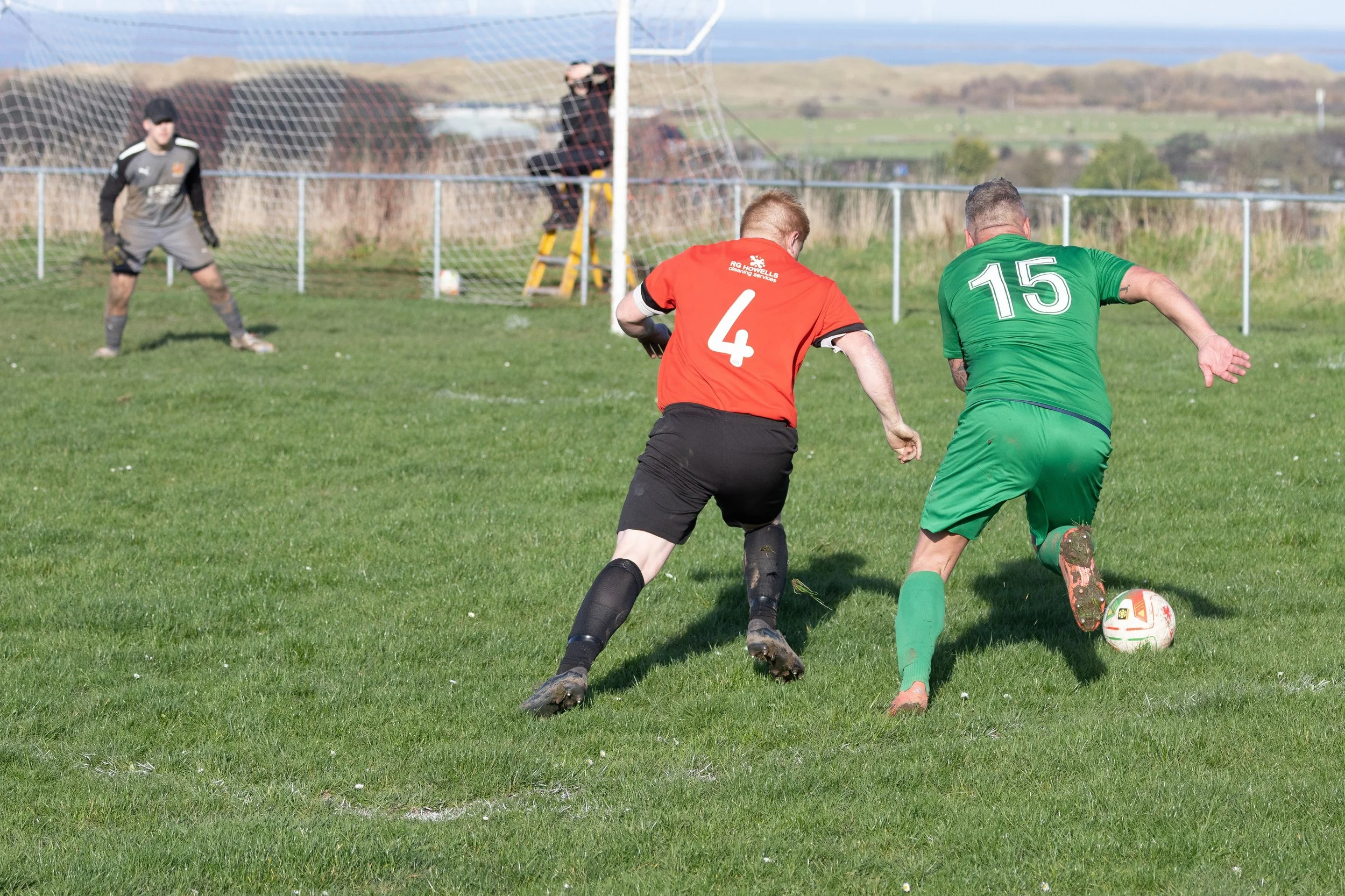 Two soccer players compete for the ball while a goalkeeper watches from the goal in the background on a grassy field.
