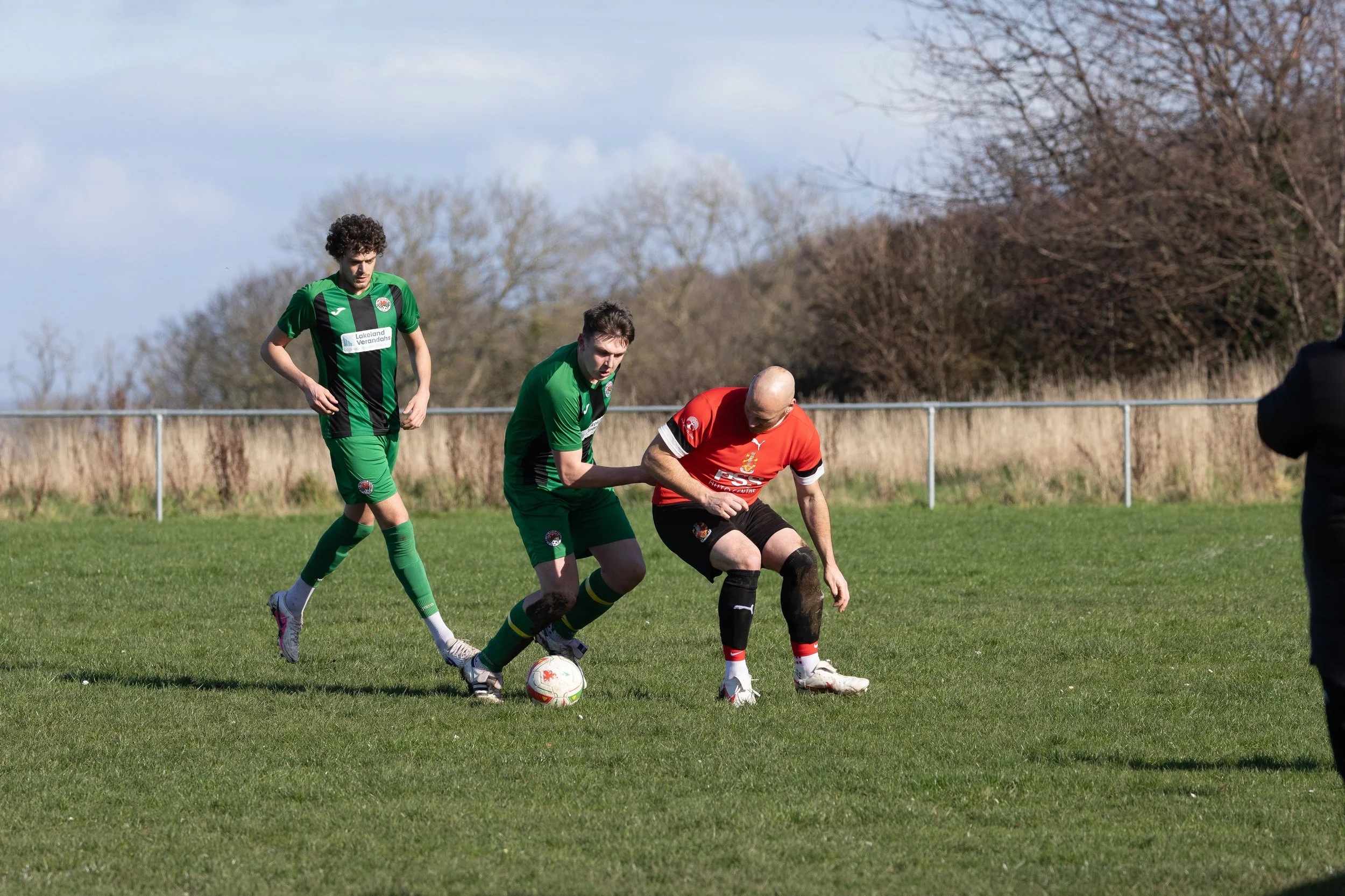 Three soccer players on a field compete for the ball, with two in green uniforms and one in a red uniform, and a person watching on the side.