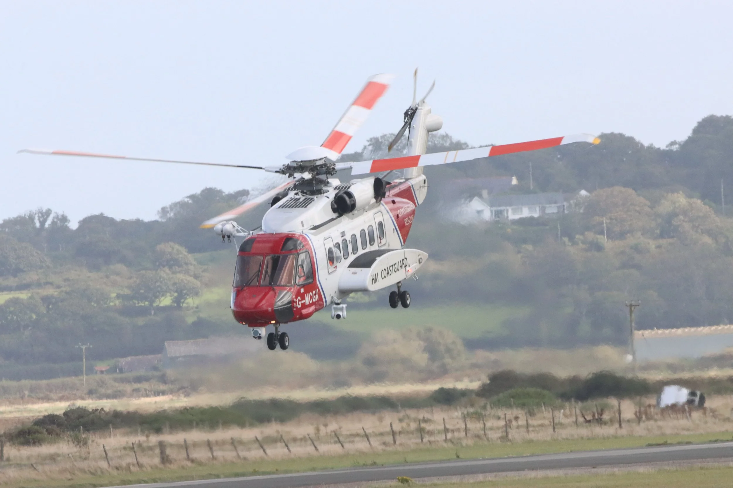 A helicopter in mid-air over a rural landscape with fields, trees, and houses in the background.