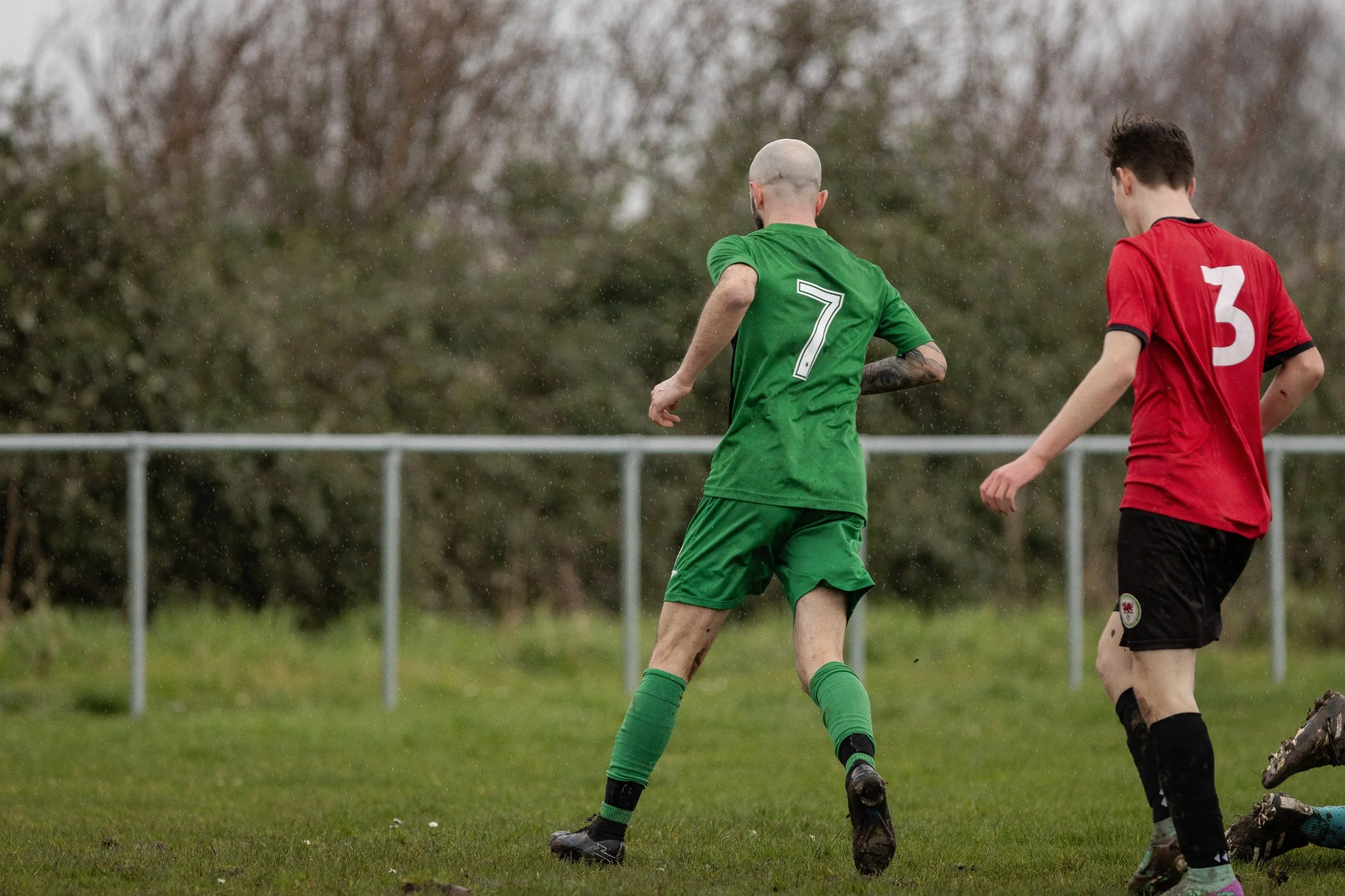 Two soccer players on a grassy field, one in a green uniform with the number 7, and the other in a red uniform with the number 3, during a match in rainy weather.