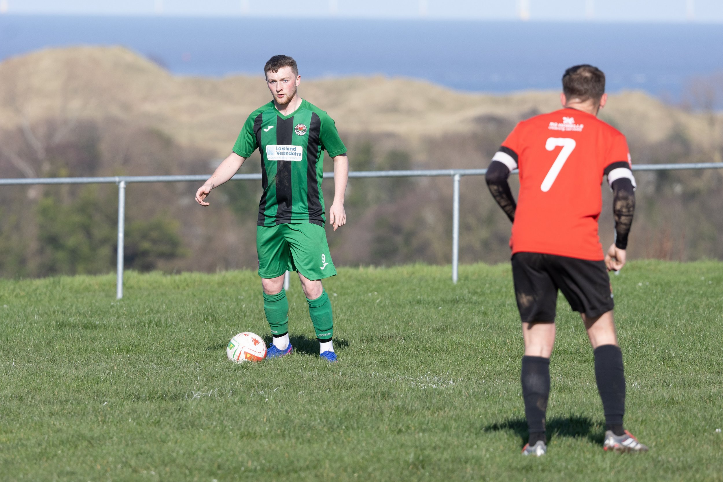 Two soccer players on a field during a match, one in a green and black uniform preparing to kick the ball, the other in a red uniform with the number 7 on the back, standing with hands on hips, in front of a railing and scenic landscape.