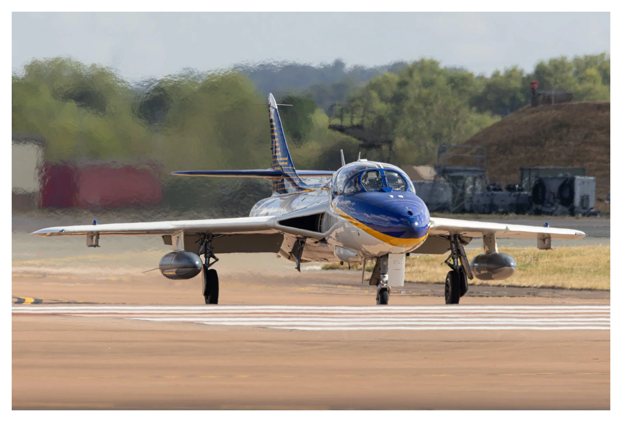 A fighter jet on a runway with a blue and white body and yellow accents, with a landscape of trees, a grassy hill, and some infrastructure in the background.
