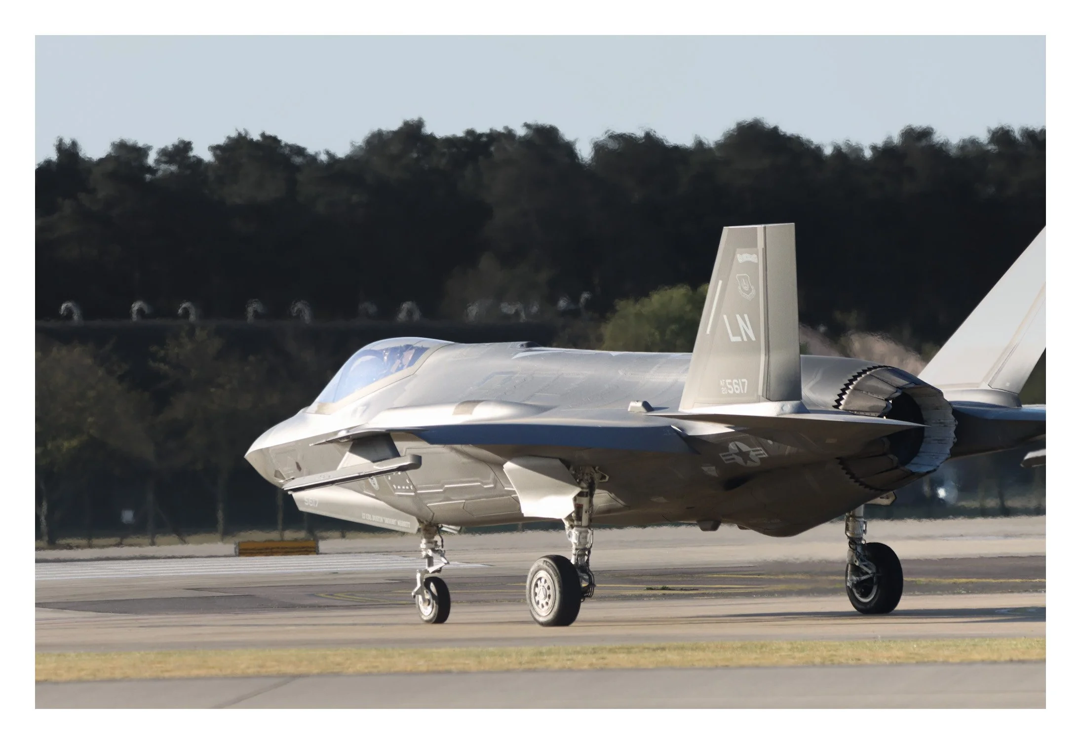 A military stealth fighter jet taxiing on a runway with trees and a blue sky in the background.