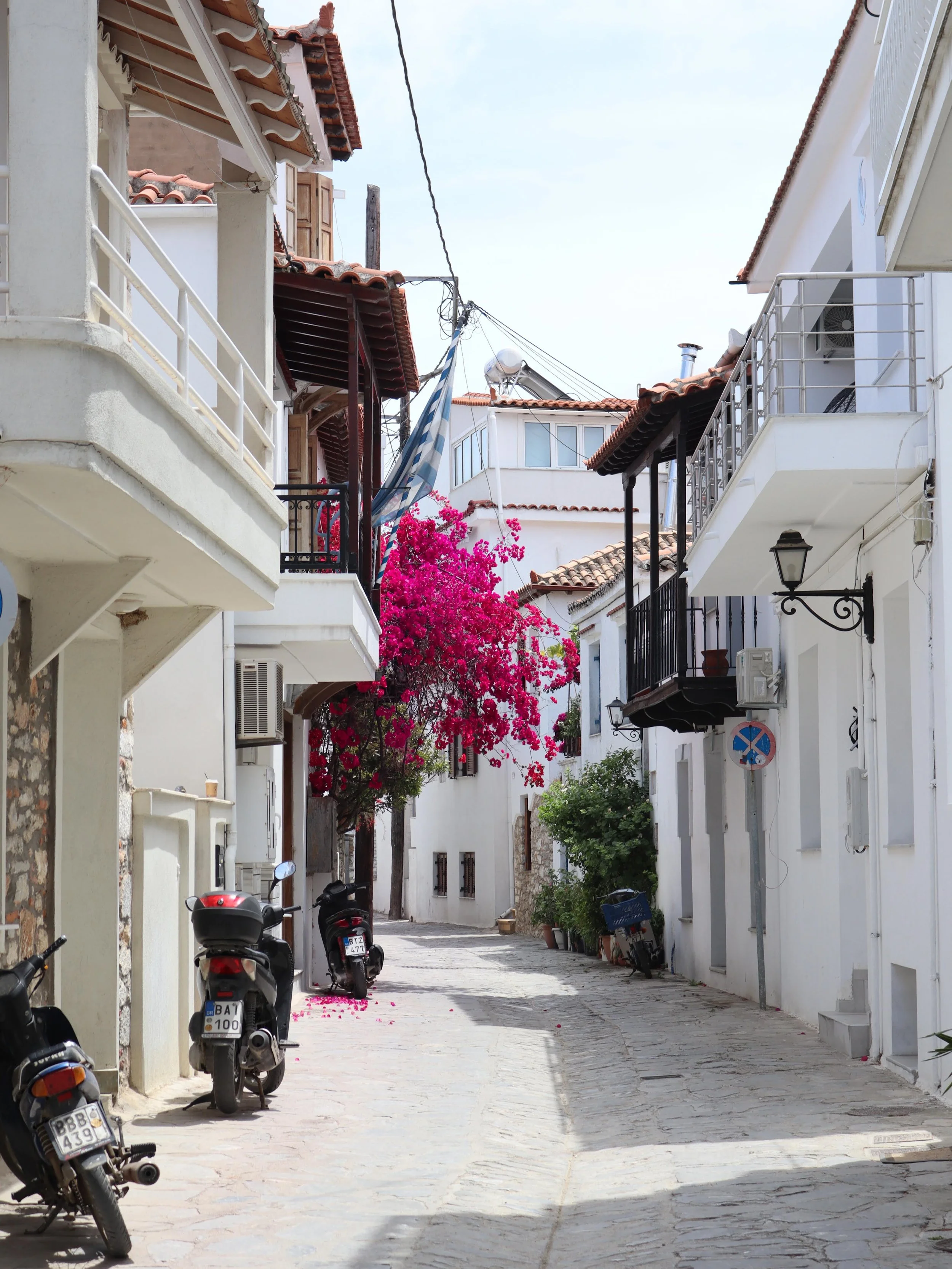 A narrow street in a Mediterranean town, lined with white buildings, some with balconies, and a flowering pink bougainvillea tree. Motorbikes are parked along the sidewalk, and power lines run overhead.