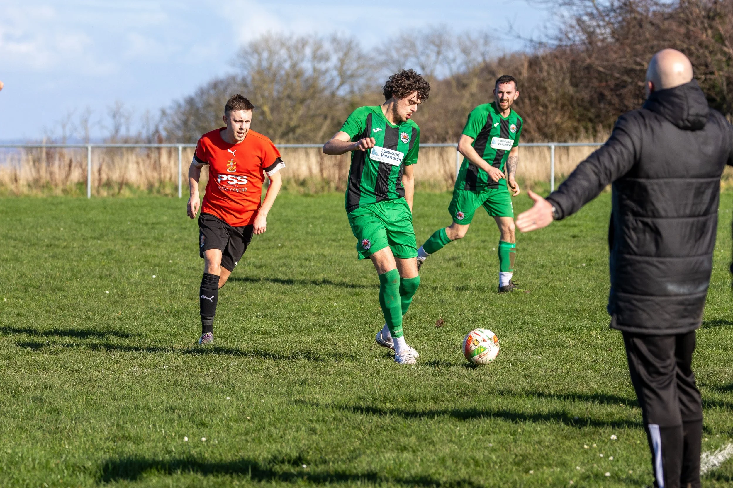 Soccer players on the field with one player in green preparing to kick the ball while being watched by an opponent in red and a coach or referee in black