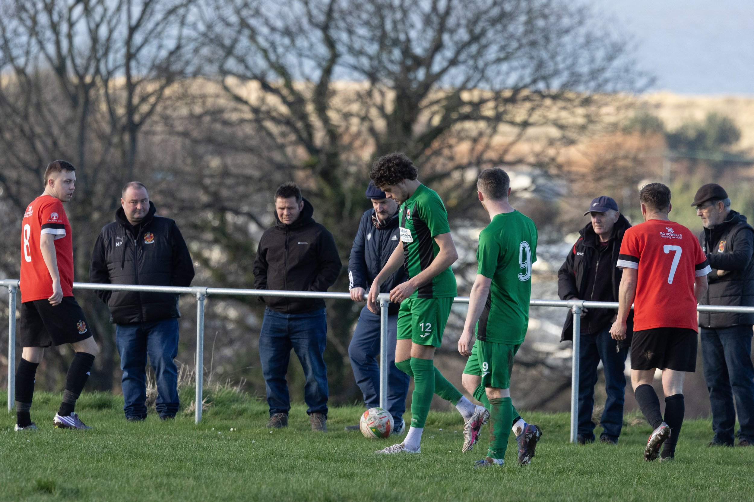 Soccer players and coaches standing by a fence on a grassy field during a match.