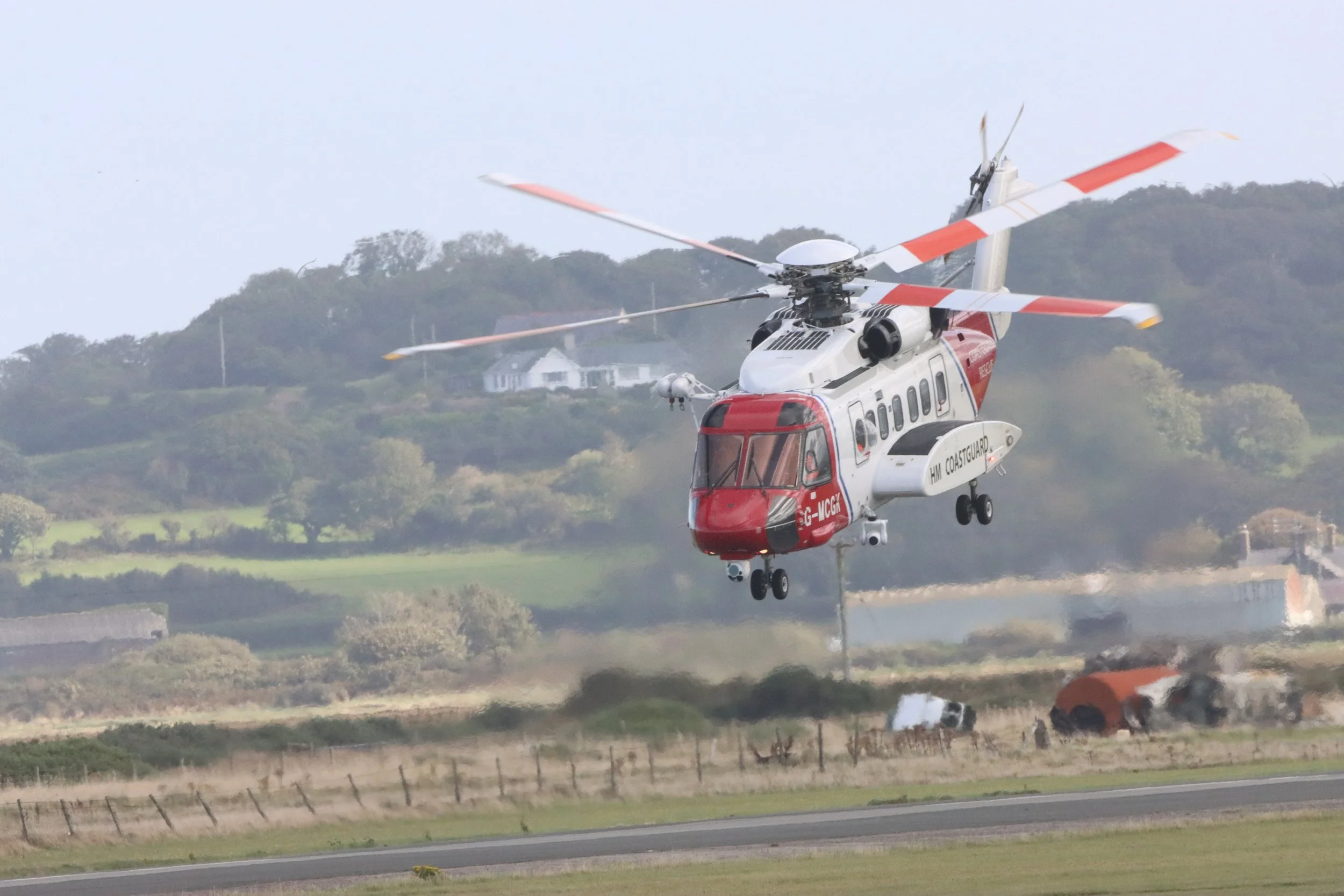 A red and white helicopter flying low over a field, with vehicles and dust visible on the ground below.