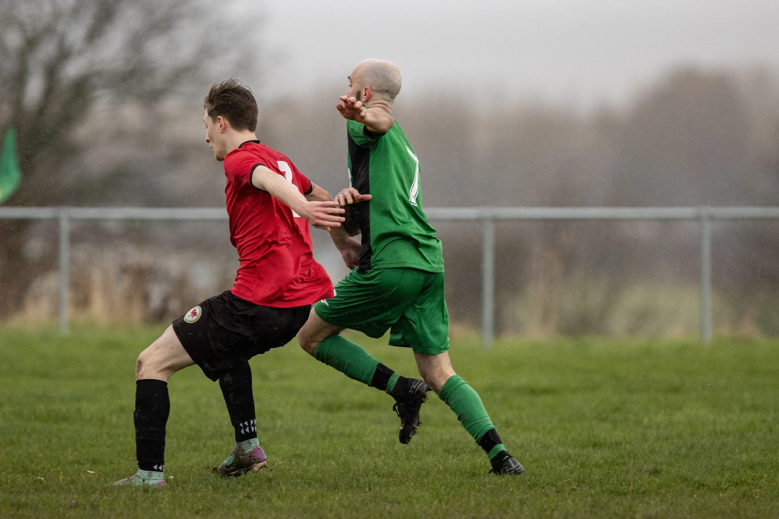 Two soccer players, one in a red shirt and black shorts and the other in green, competing for the ball on a grassy field in rainy weather.