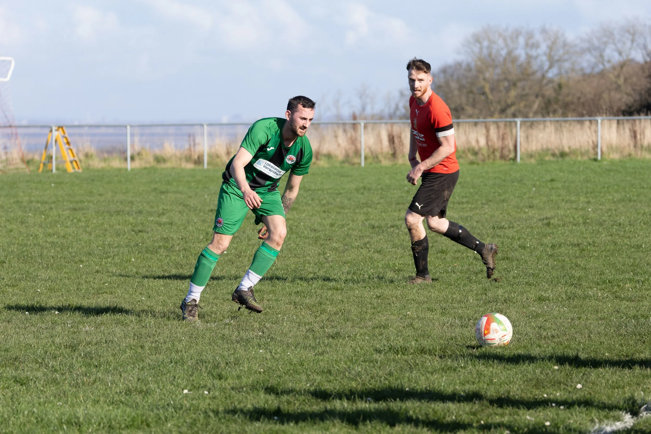 Two soccer players on a grassy field with a soccer ball between them, one in a green uniform and the other in a red and black uniform, during a match.