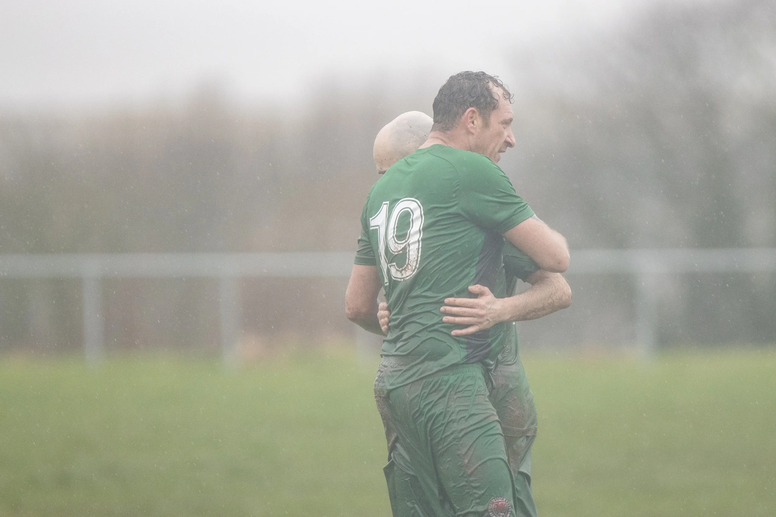 Two soccer players in green uniforms standing on a rainy field, embracing each other, with a blurred background of a fence and trees.