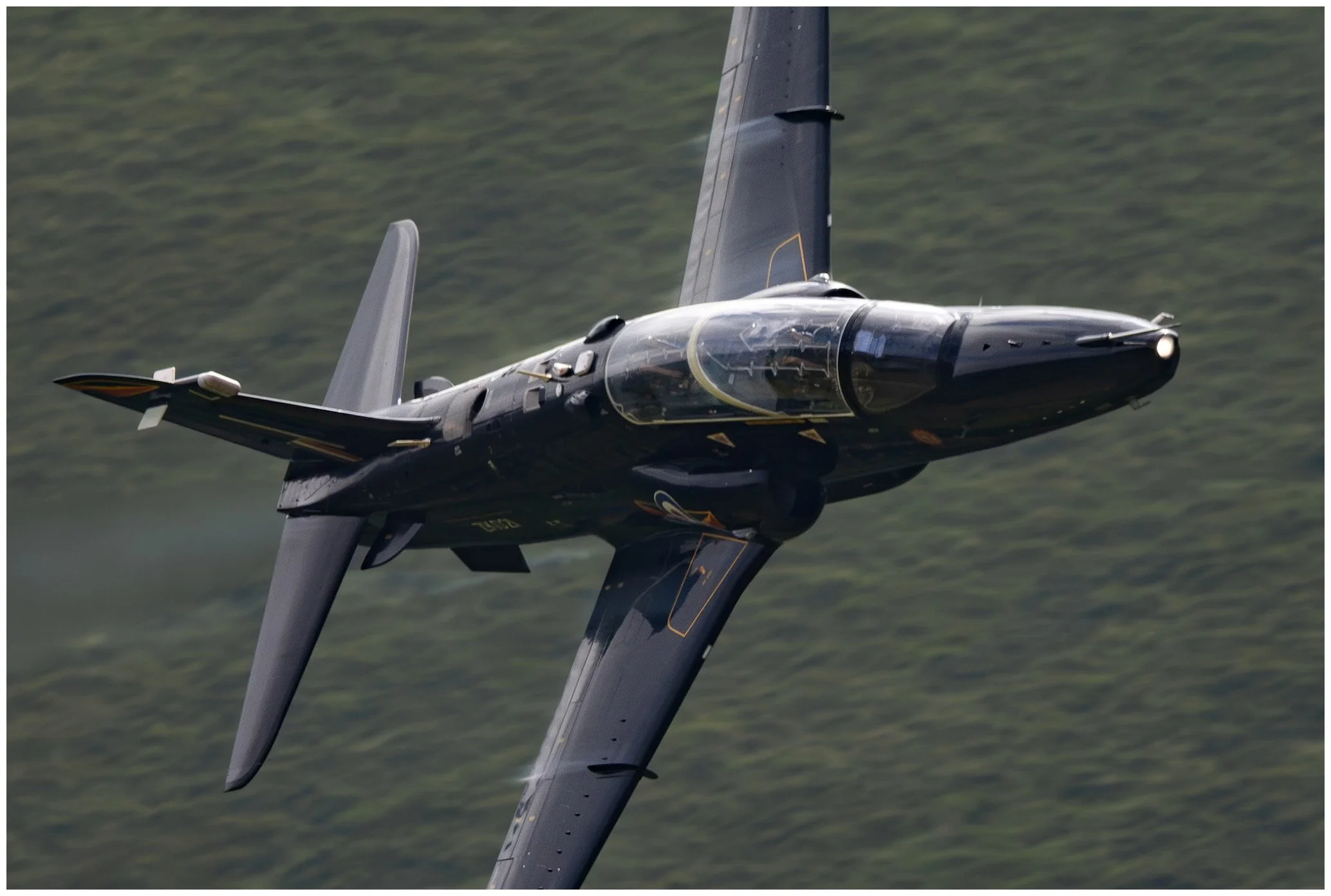 A fighter jet flying over a landscape with trees, seen from below and tilted at an angle.