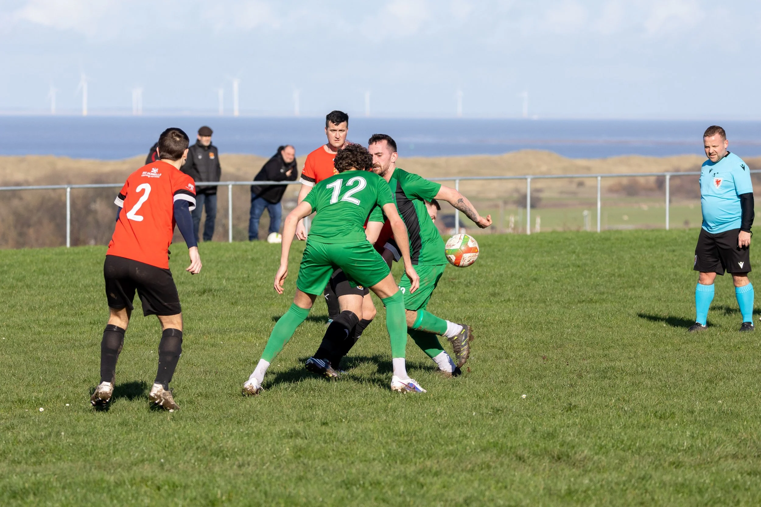 Soccer players in green and red uniforms competing for the ball on a grassy field with a referee observing in the background.