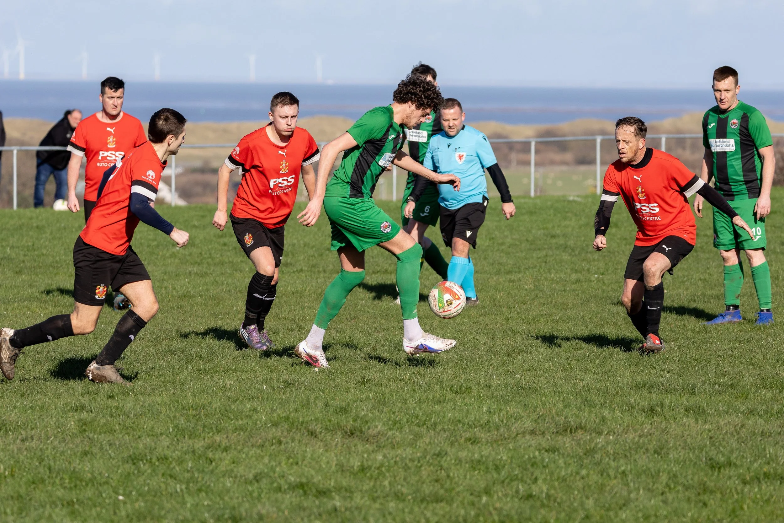 A group of soccer players in red and green jerseys and black shorts are playing on a grassy field, with some spectators and wind turbines in the background.