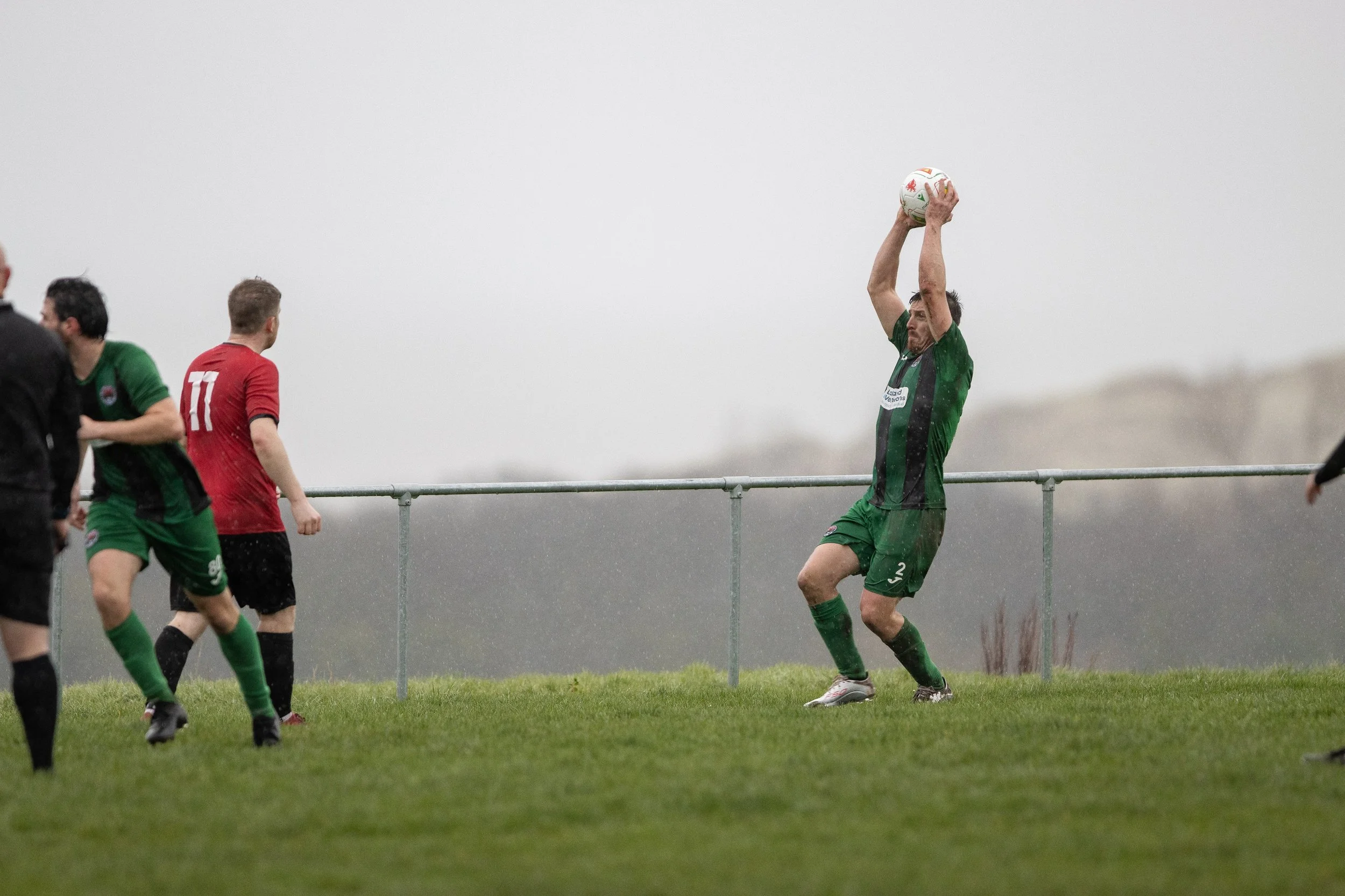 A soccer player in a green and black uniform preparing to throw in a match in rainy weather, with other players nearby on the field.