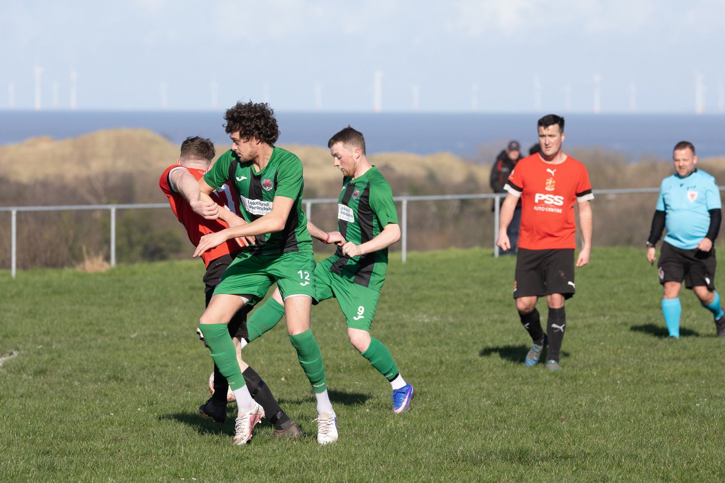 Soccer match with players competing for the ball on a grassy field, with a light blue sky and wind turbines in the background.