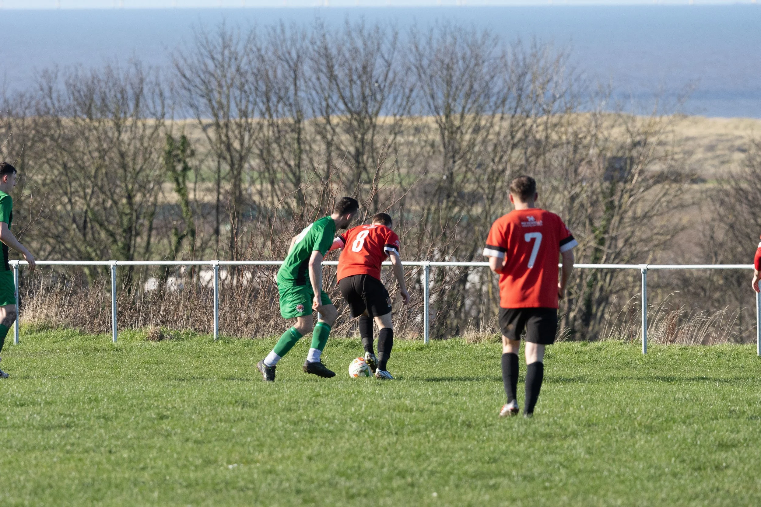 Soccer players in green and red jerseys competing for the ball on a grassy field, with a background of leafless trees and a fence.