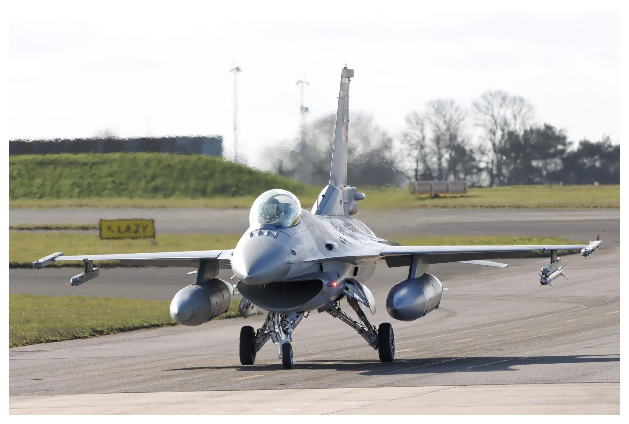 A military fighter jet taxiing on an airstrip, equipped with missiles and fuel tanks, with a background of trees and a cloudy sky.