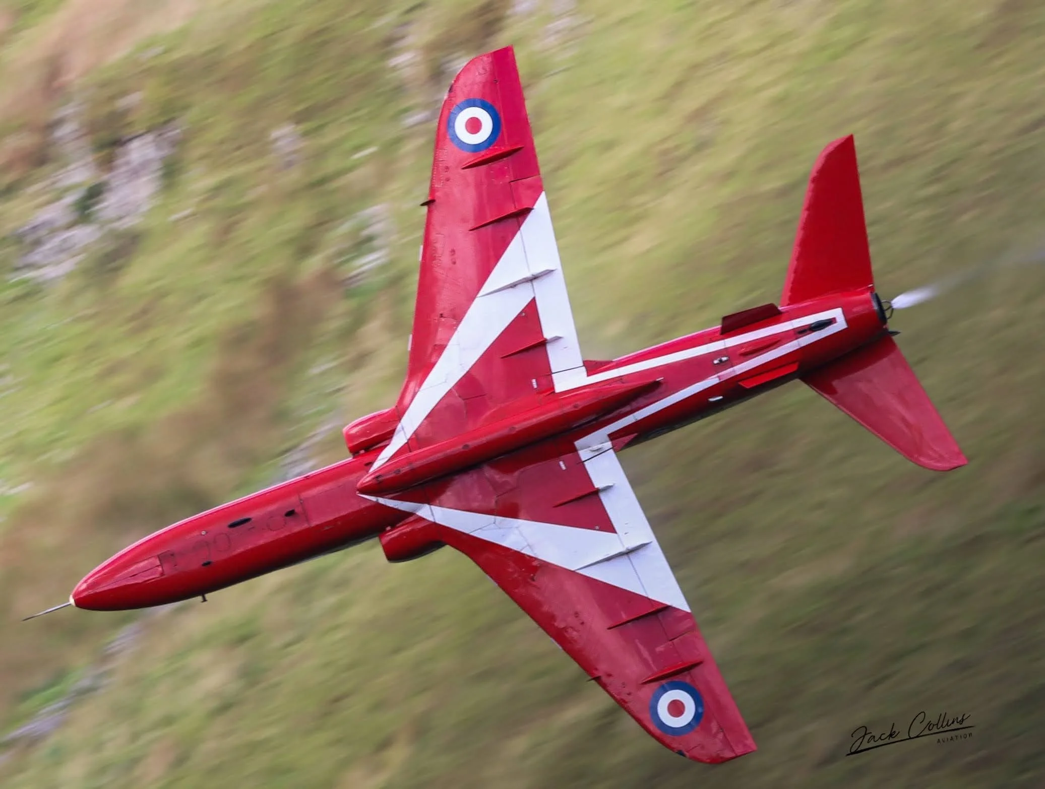 Red and white jet airplane in flight over grassy landscape, with the British Royal Air Force roundels on the wings.