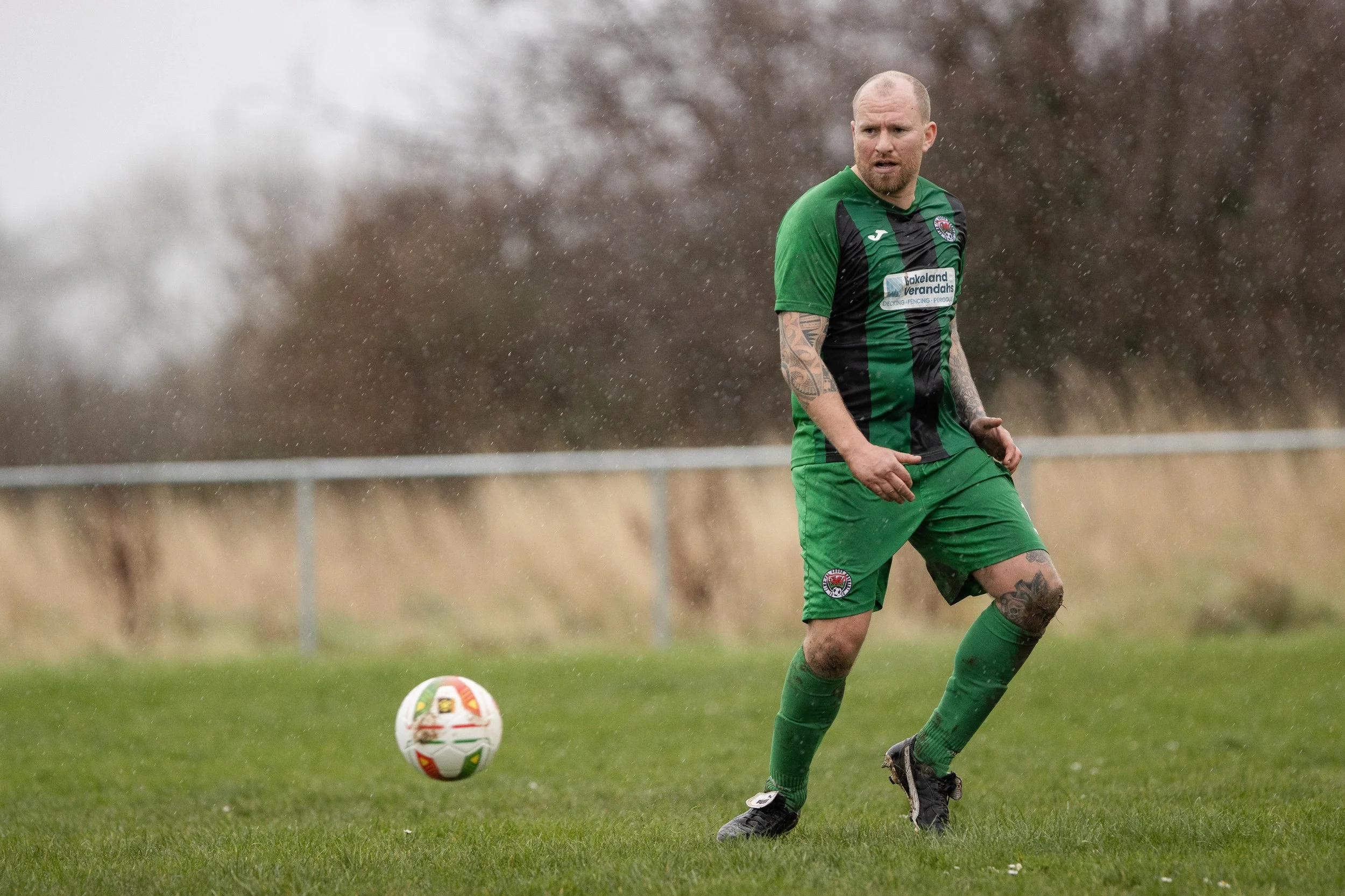 A man in a green and black soccer uniform playing soccer on a rainy field, with a ball nearby and trees in the background.